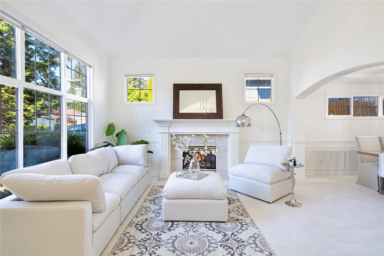 Bright living room with white sofa, chair, and ottoman arranged around a fireplace with a mirror above. Large windows let in natural light.