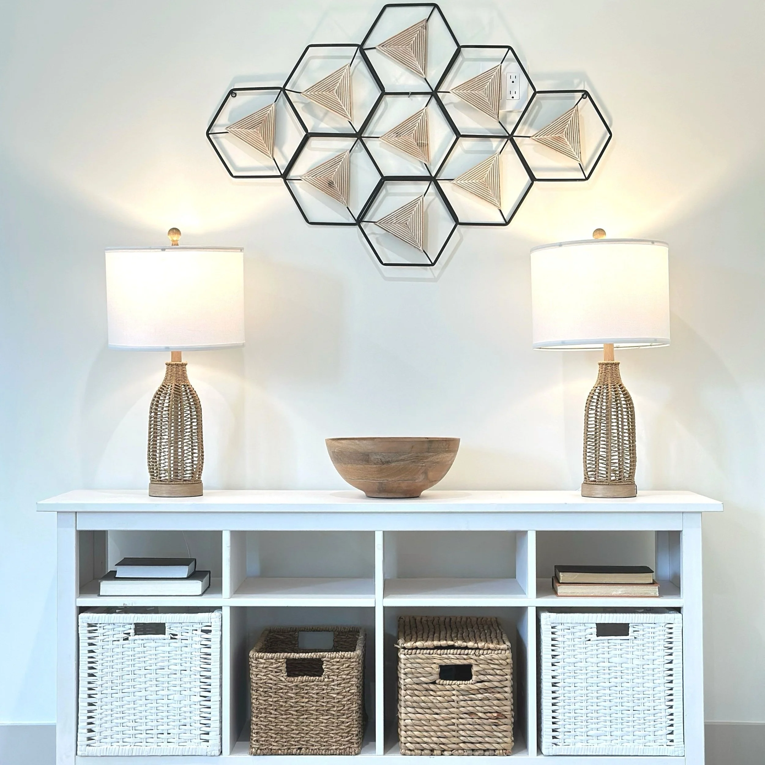 Decorative wall art made from black metal honeycomb frames with woven wooden elements, flanked by two table lamps with wicker bases and white shades on a white console table with baskets and books underneath.