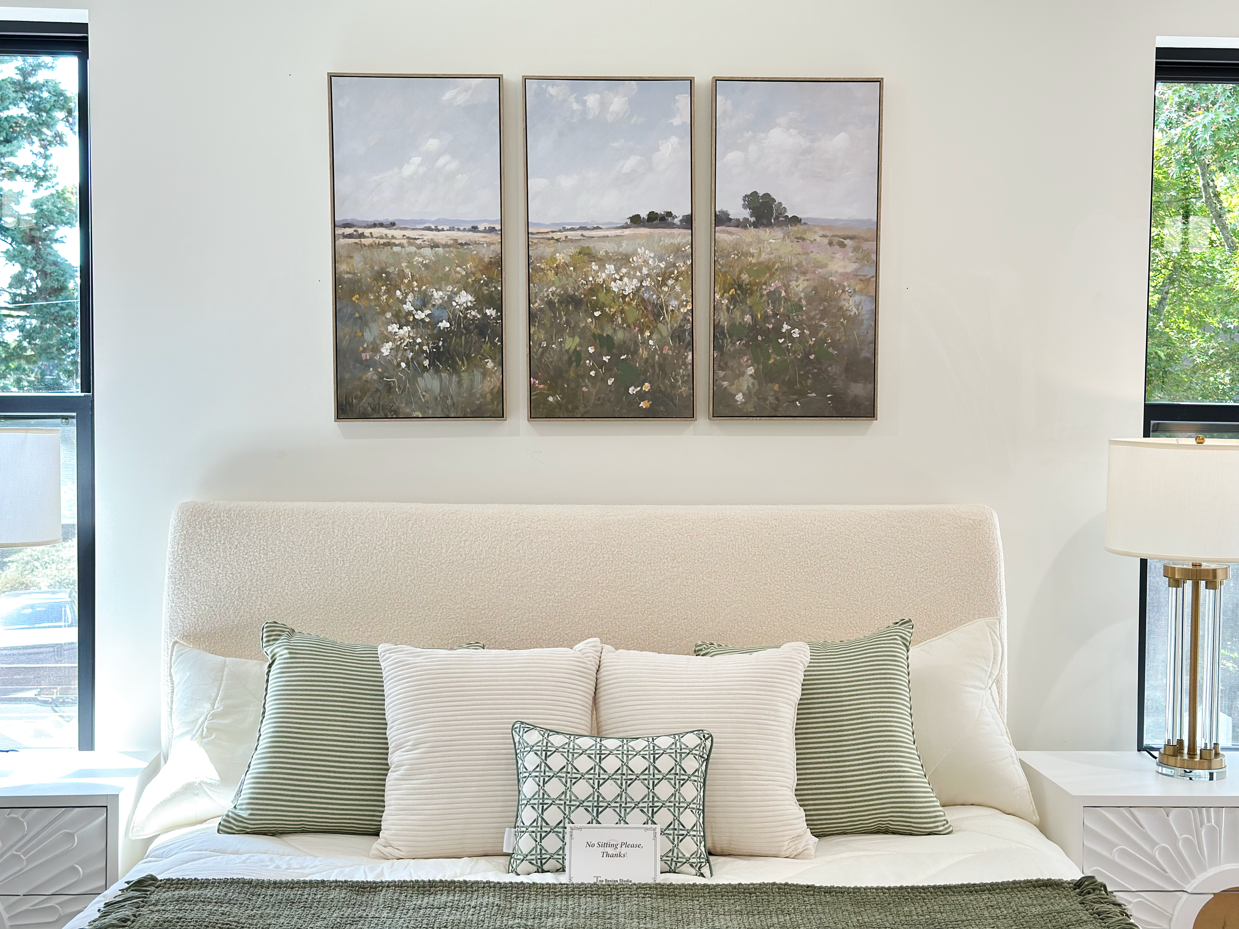 A bedroom with a white bed, multiple pillows, side tables with lamps, and three framed landscape paintings on the wall. There are windows on both sides of the room showing outside greenery.