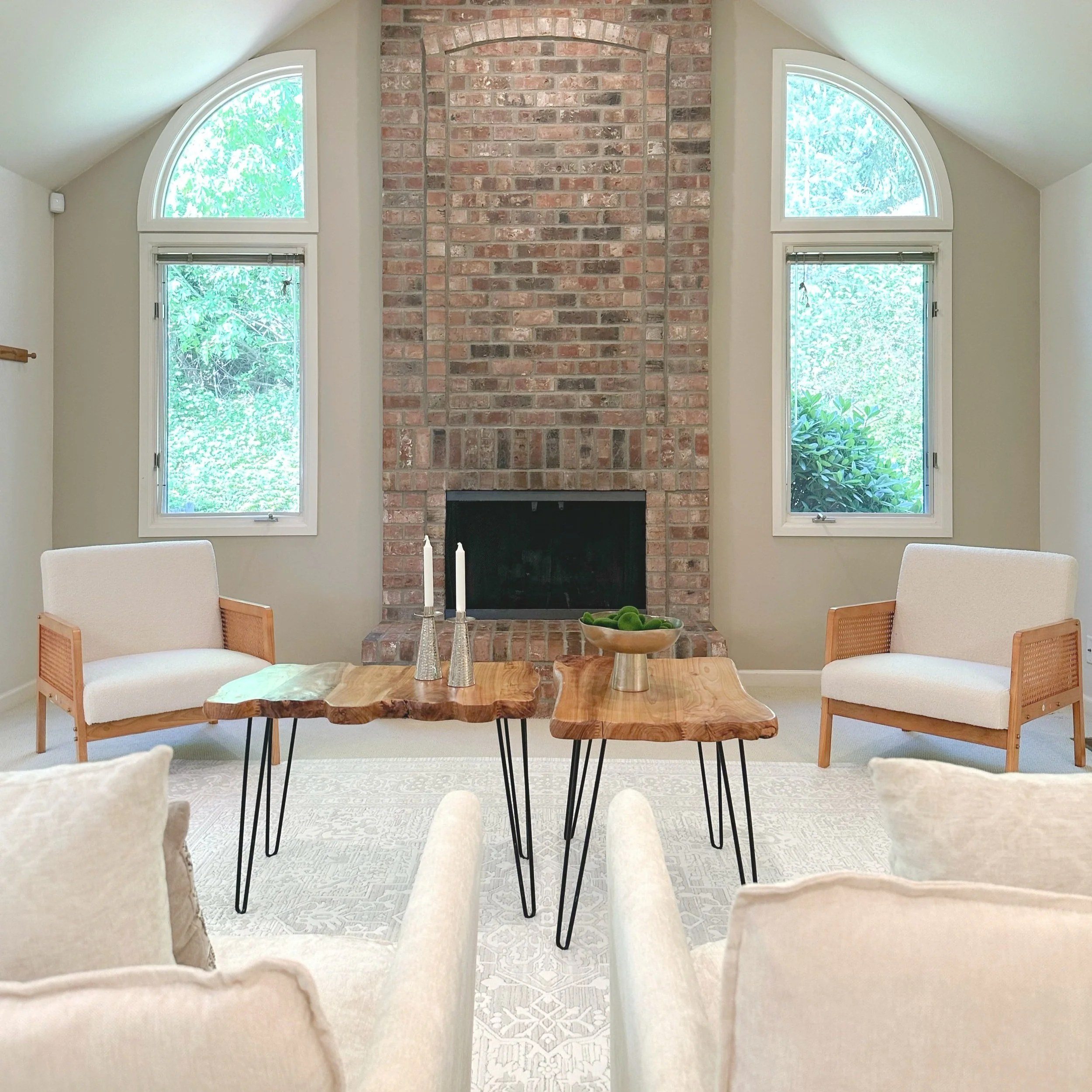Living room with brick fireplace, two arched windows, beige walls, cream-colored armchairs, two rustic wooden coffee tables with black metal legs, and decorative candles and a bowl on the tables.
