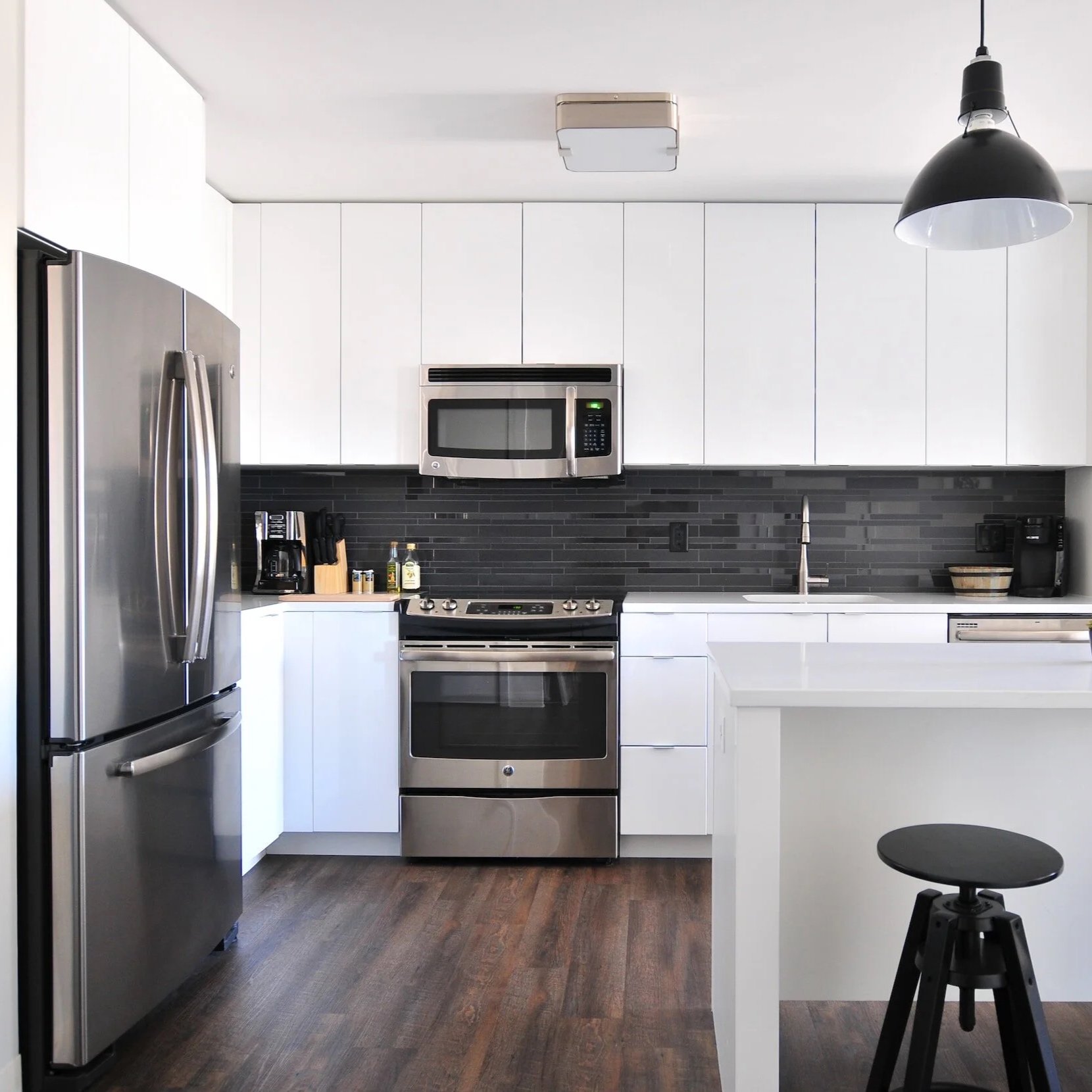 Modern kitchen with white cabinets, black backsplash, stainless steel appliances including refrigerator, microwave, and oven, black pendant light, black stool, and dark wood flooring.