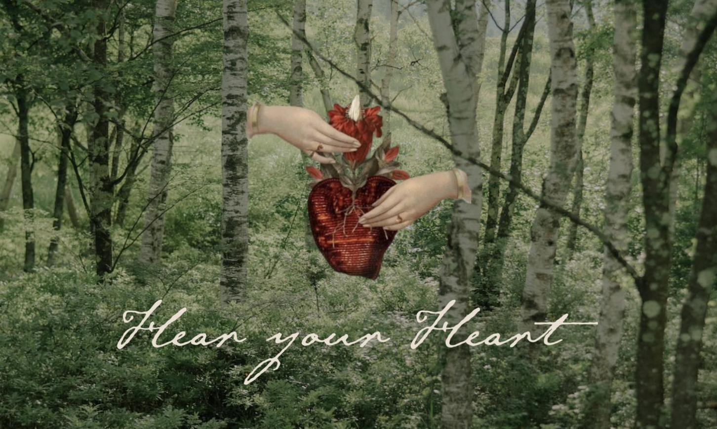 A digitally manipulated image of a forest scene with two hands holding a bleeding red heart, with a flower and a unicorn horn on a top of the heart, and the text 'Hear your Heart' written in cursive at the bottom.