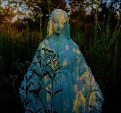 Sculpture of a woman with a veil, painted with vibrant colors, surrounded by plants in an outdoor setting at sunset.