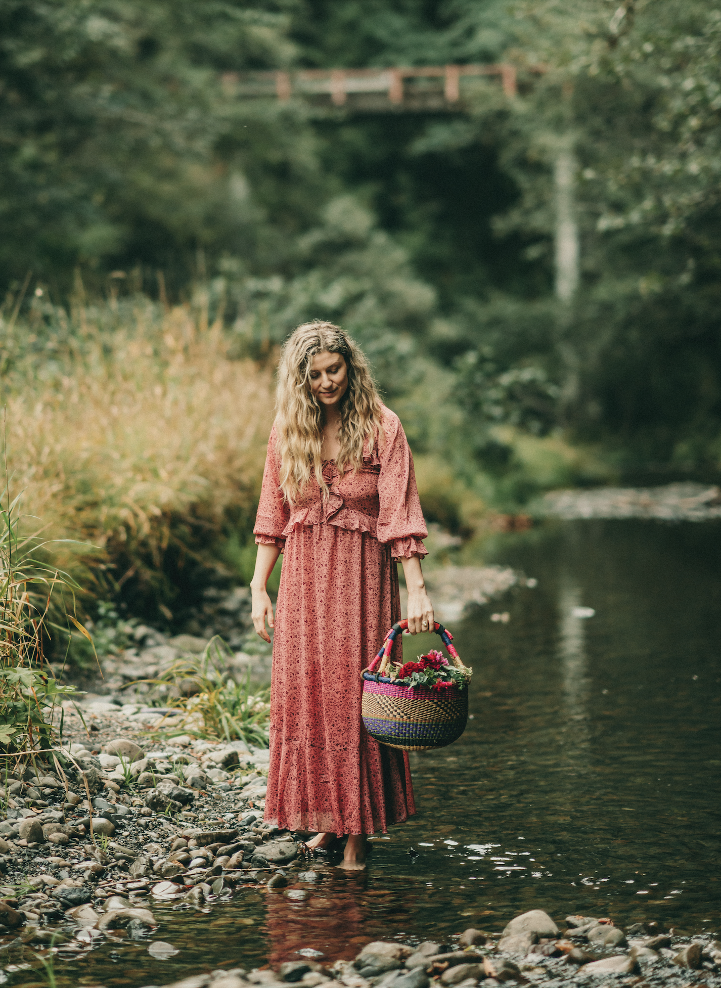 A woman in a long pink dress standing barefoot in a shallow river, holding a colorful woven basket filled with flowers, surrounded by green trees and rocks.