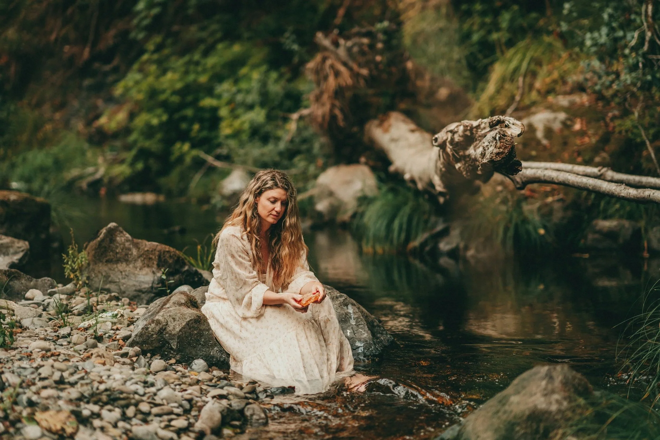 A woman in a white dress sitting on rocks by a river, holding a piece of bread, surrounded by lush green foliage.