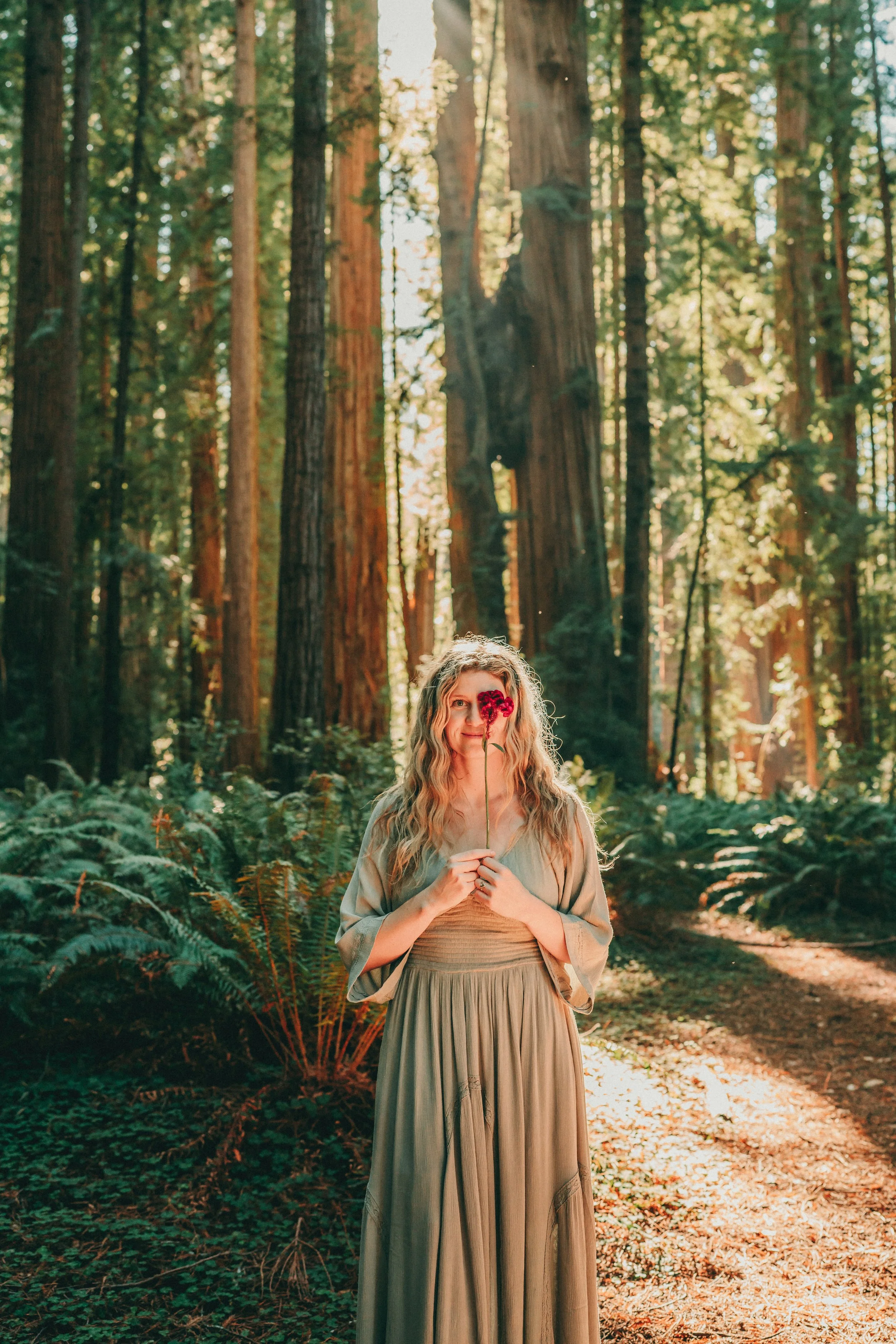 A woman with long, curly blonde hair standing in a forest with tall trees and green foliage. She is holding a pink flower close to her face, partially covering one eye, and is smiling.
