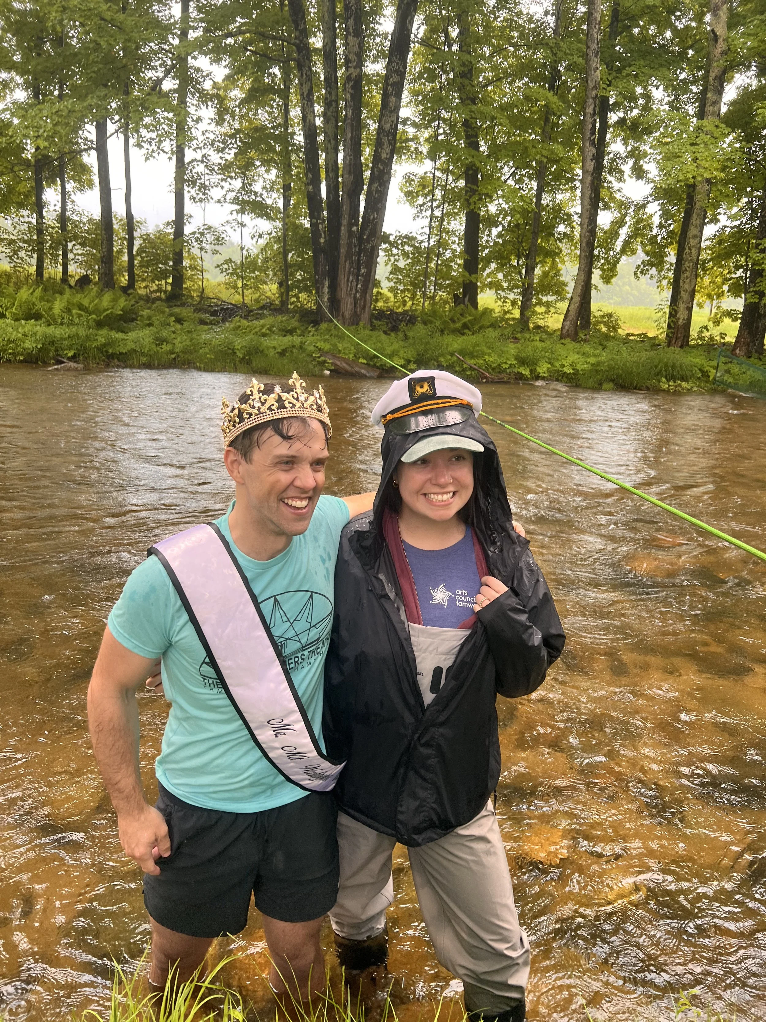 Man in a crown and sash standing next to a woman in a boat captain's hat, both in a river.