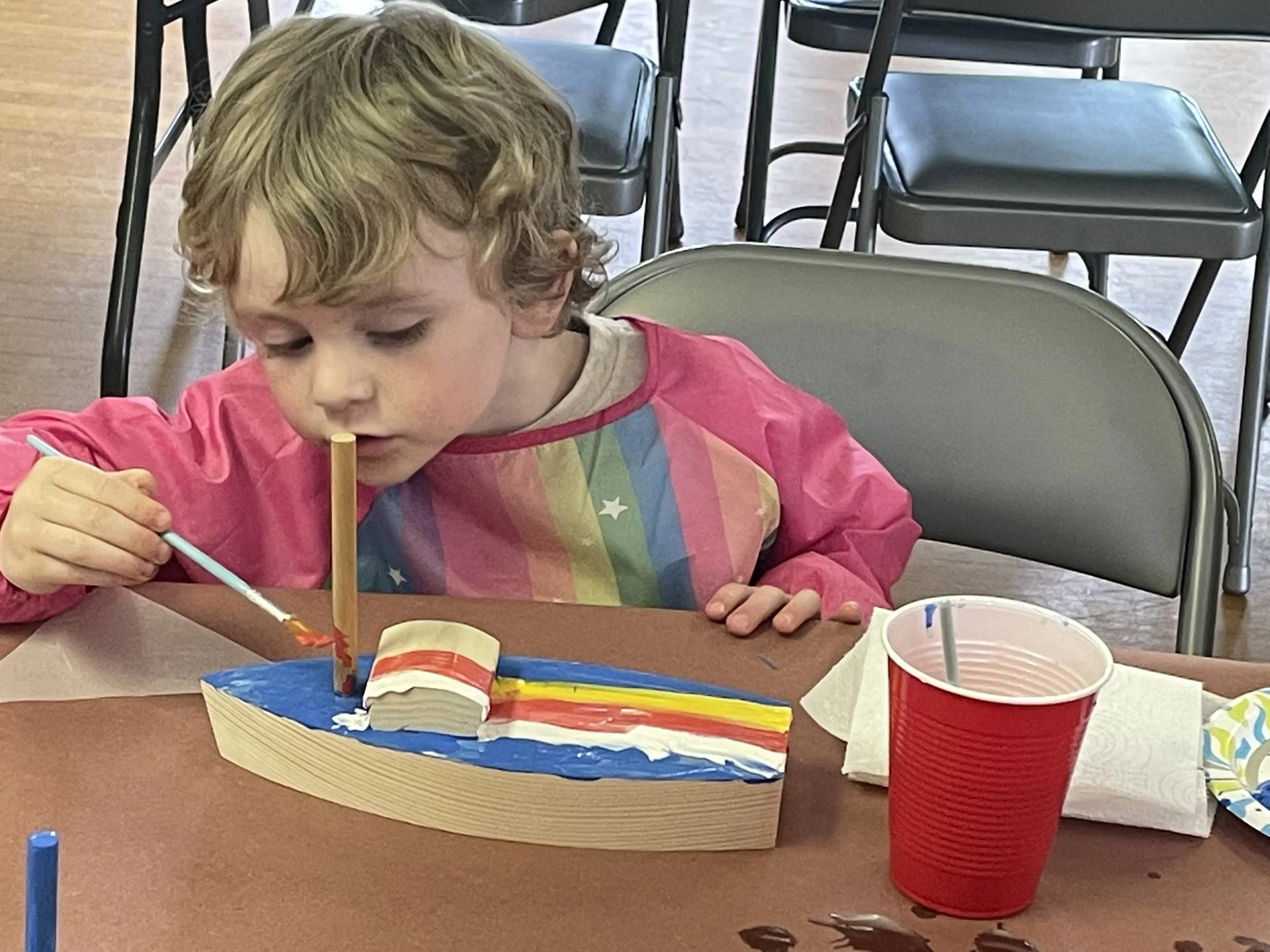 A young child is painting a colorful wooden boat at a table, using a paintbrush and surrounded by crafting materials, including a red plastic cup.