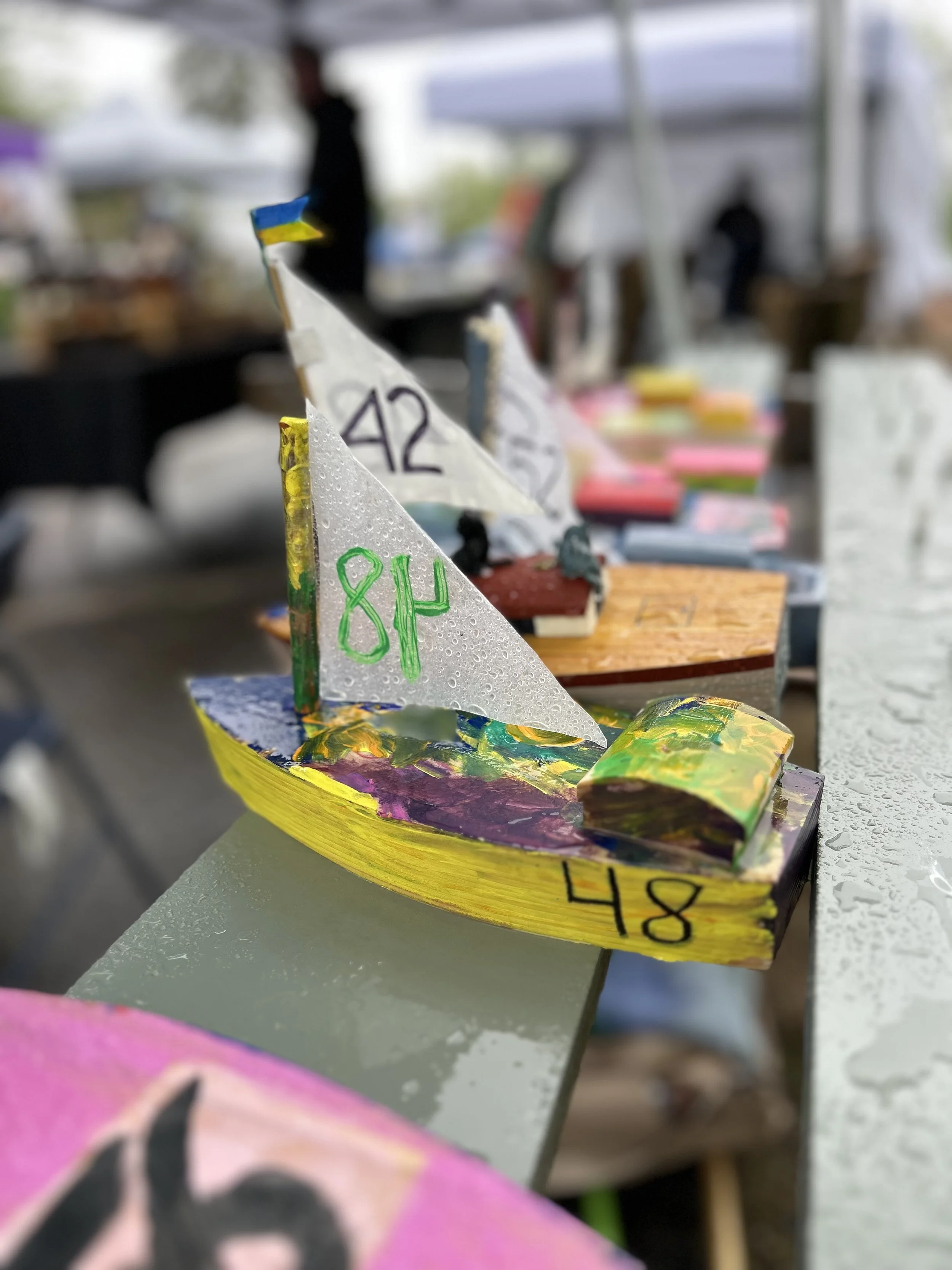 A close-up of a small, colorful model sailboat with the number 84 on its sail, positioned alongside other similar boats in a row. The boats are wet from rain, and there's a blurred background of more boats and people beneath a tent.