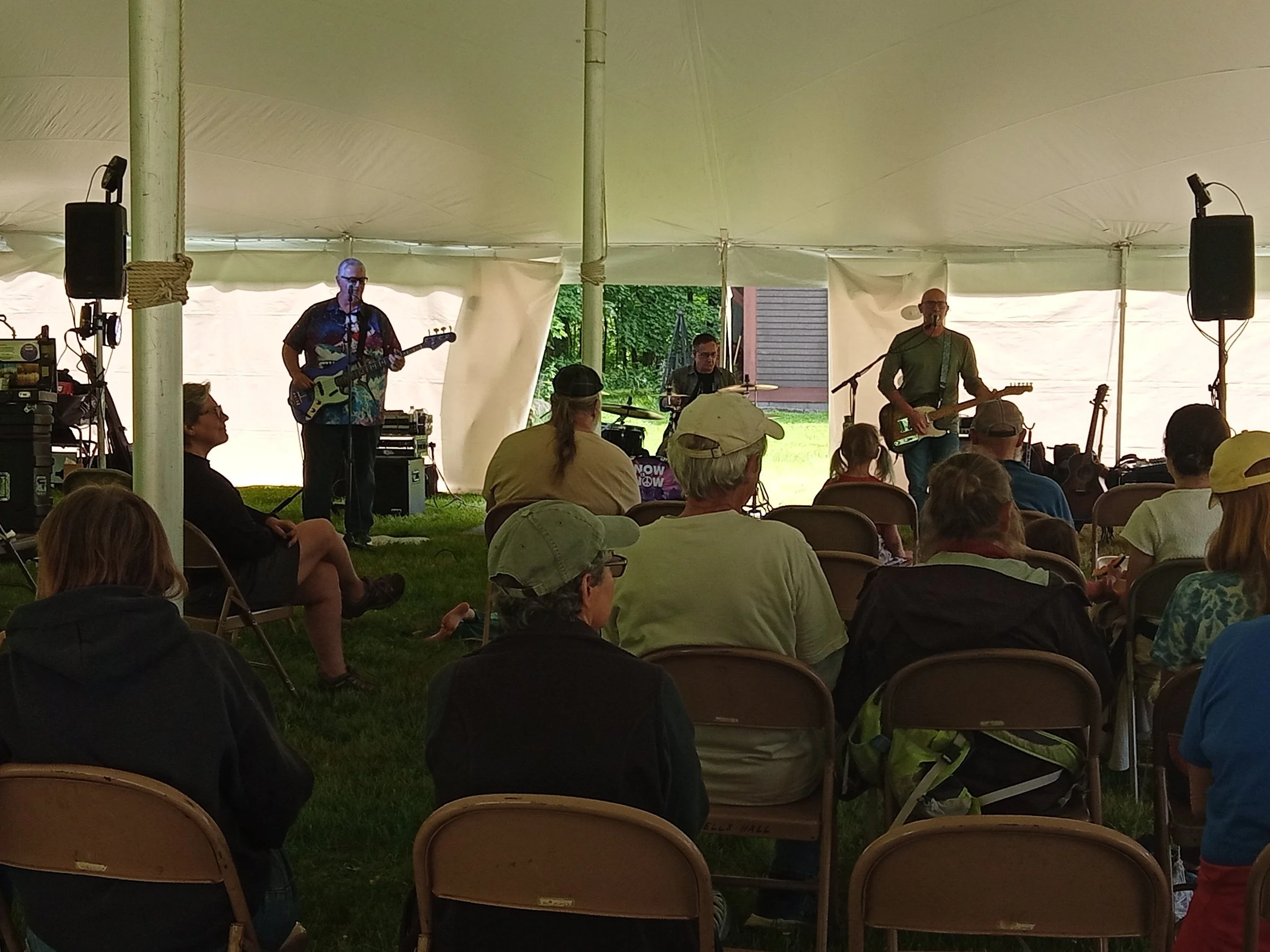 Three-person rock band playing outside under a tent in front of a seated audience.
