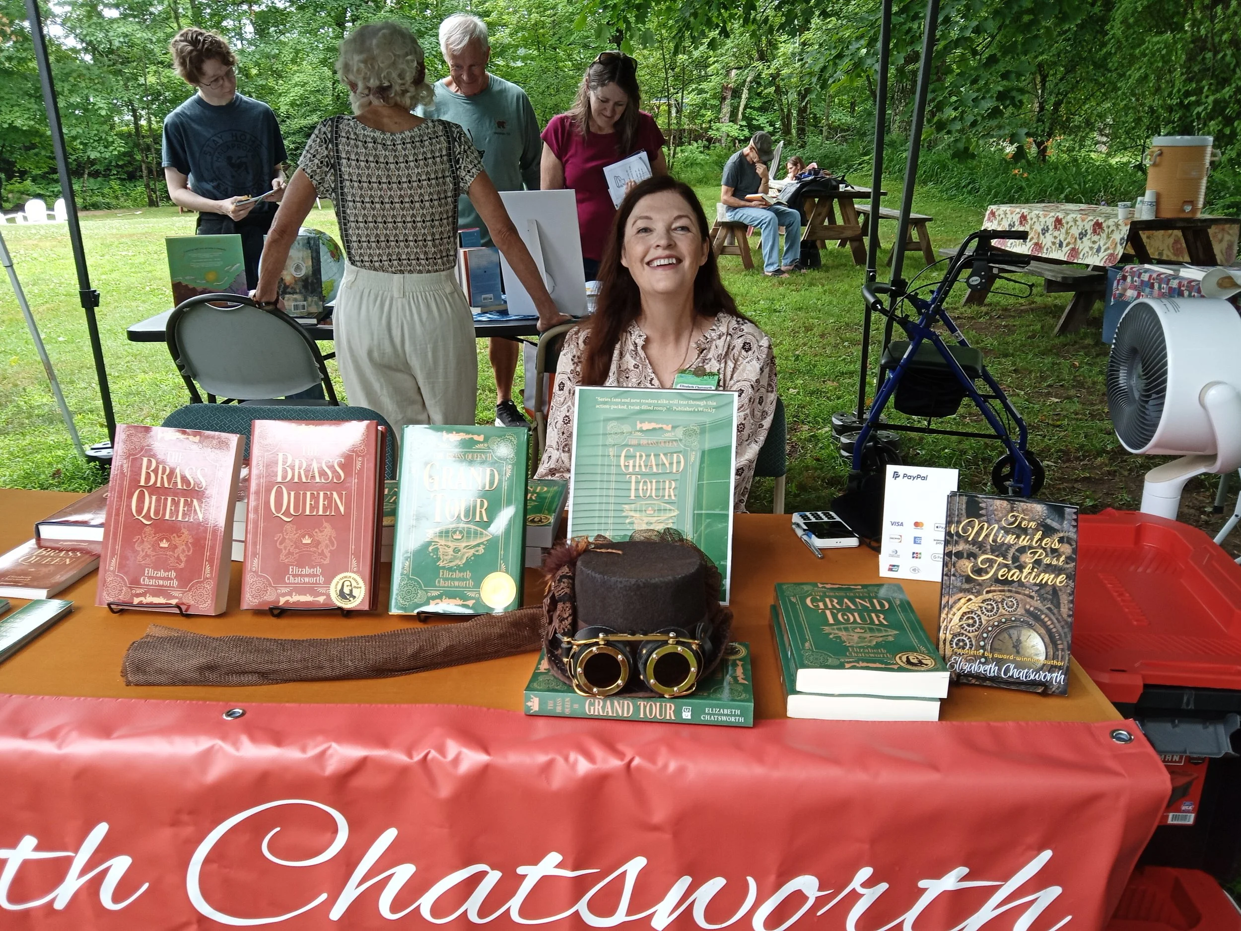 Image of a smiling woman standing behind a table filled with books and a steampunk top hat.