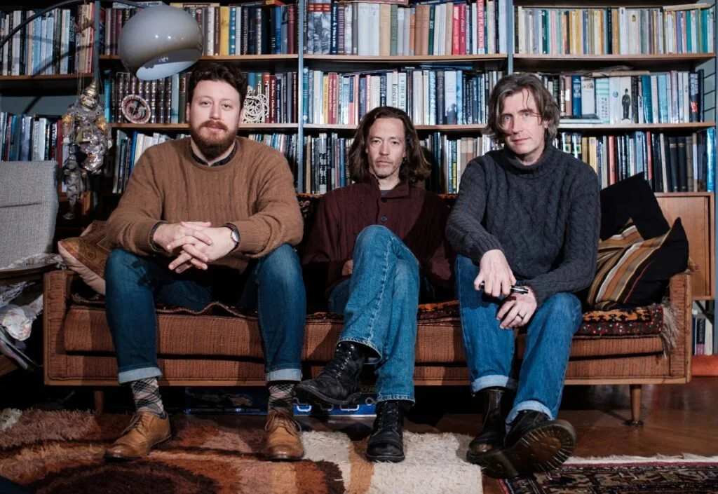 Three men sitting on a sofa in a cozy room filled with bookshelves. The man on the left has a beard and is wearing a brown sweater, the middle man has long hair and is in a dark shirt, while the man on the right has medium-length hair.