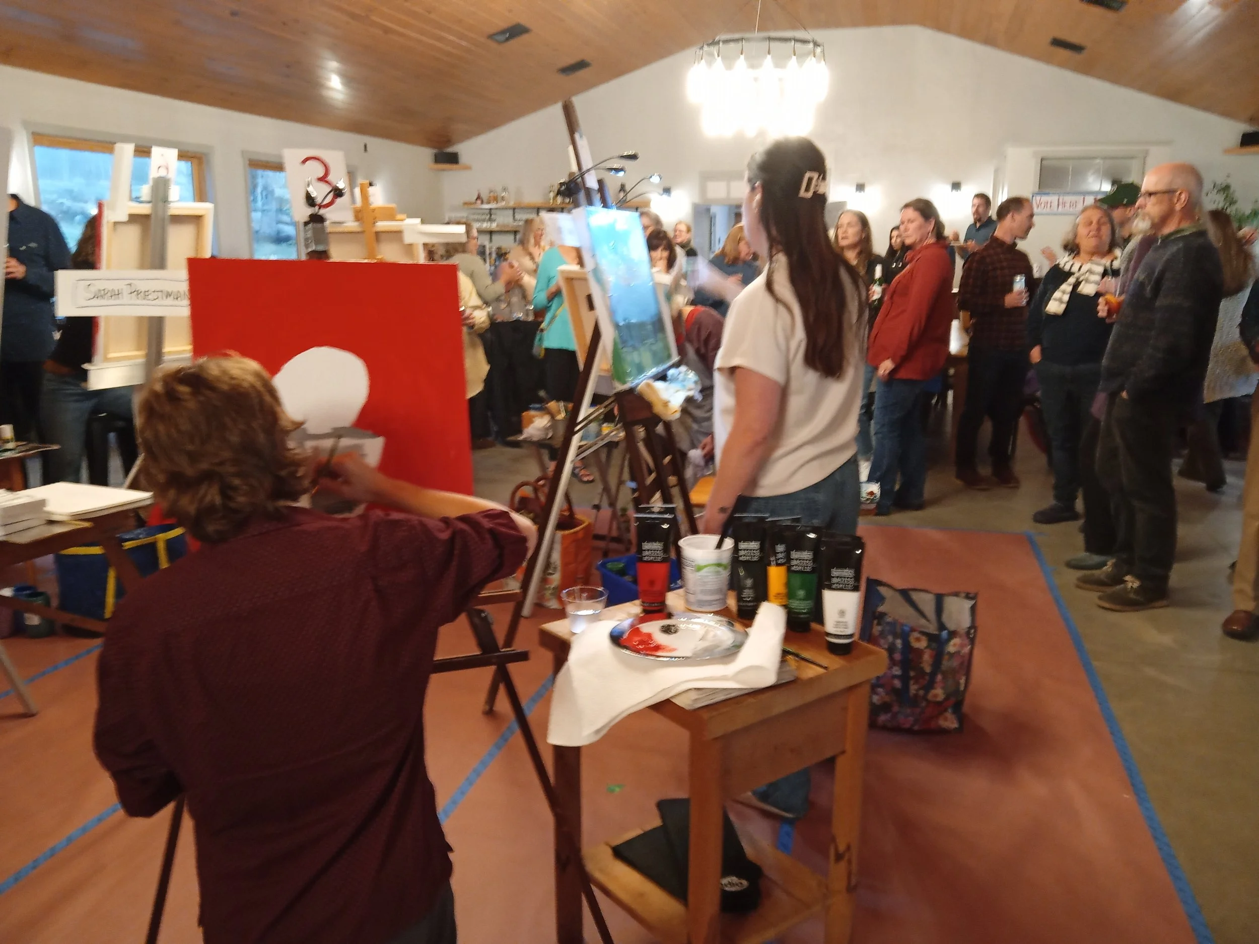 A crowd of people watching an artist paint in a spacious, well-lit room with easels and drinks on tables.