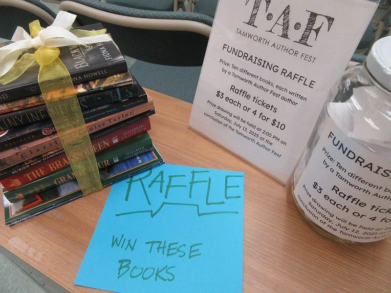 A stack of books tied with a ribbon next to a sign for a fundraising raffle at the Tamworth Author Fest, with another sign detailing raffle ticket prices. A bright blue sheet of paper on the table says "RAFFLE" and "WIN THESE BOOKS."