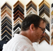 A man with short brown hair and glasses stands in front of rows of picture frames.