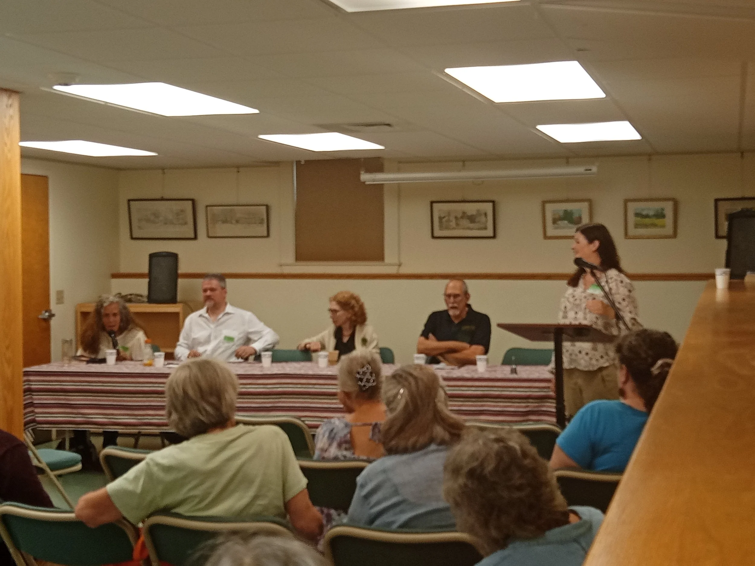 A panel discussion taking place in a small room with several people seated at a table and an audience in the foreground.