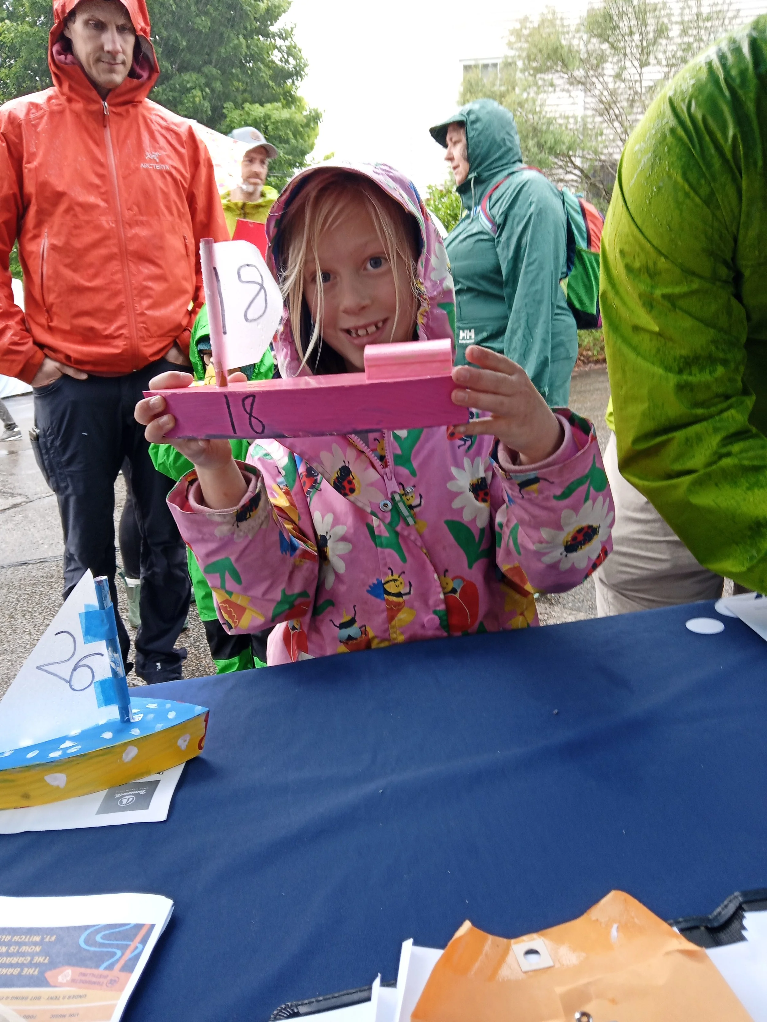 Girl in a pink raincoat holding up a wooden boat painted pink.