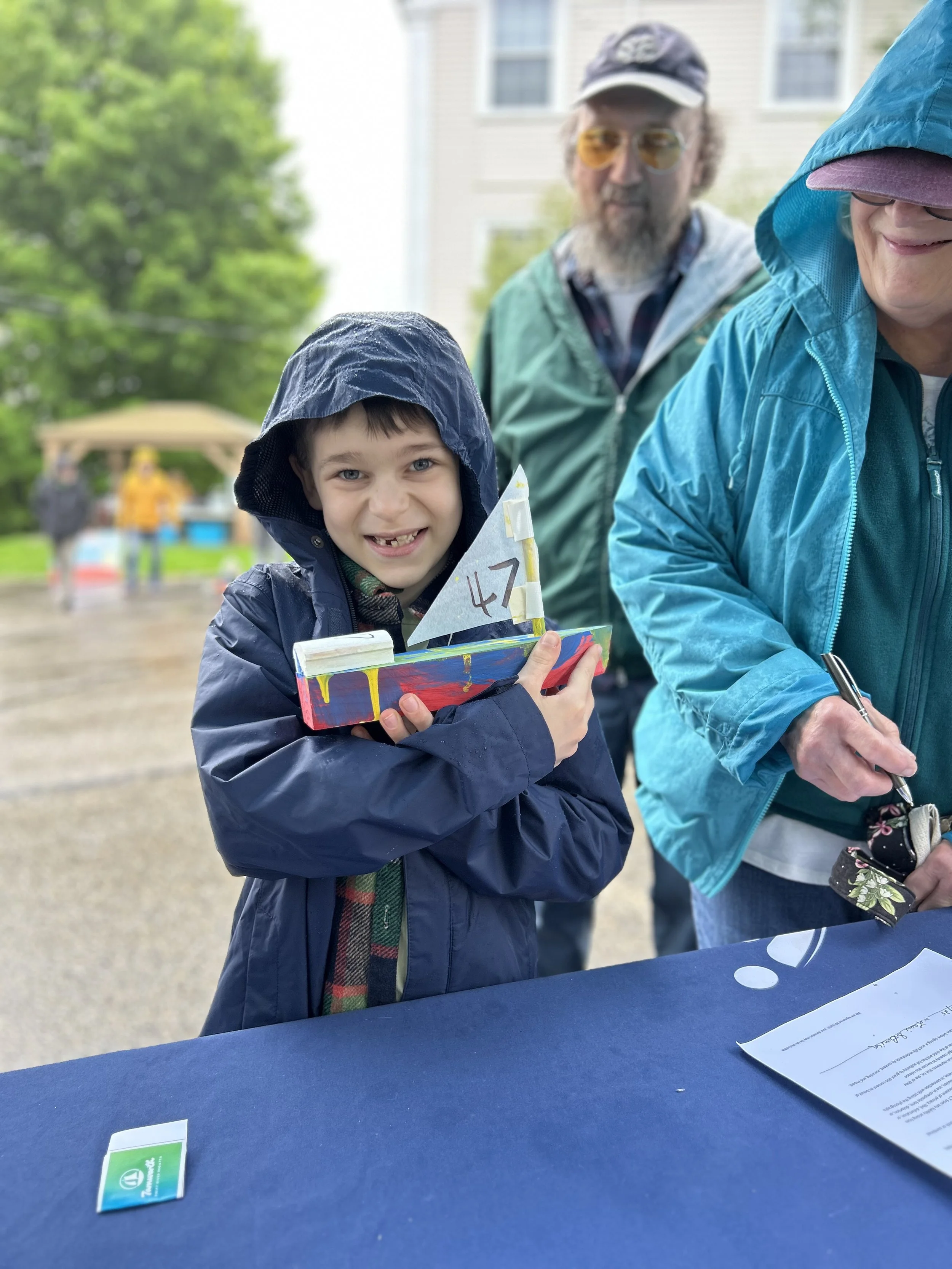 A child wearing a raincoat holds a model boat, smiling at the camera. In the background, two adults are partially visible, one is wearing glasses and another is writing on a clipboard. It appears to be an outdoor event in rainy weather.