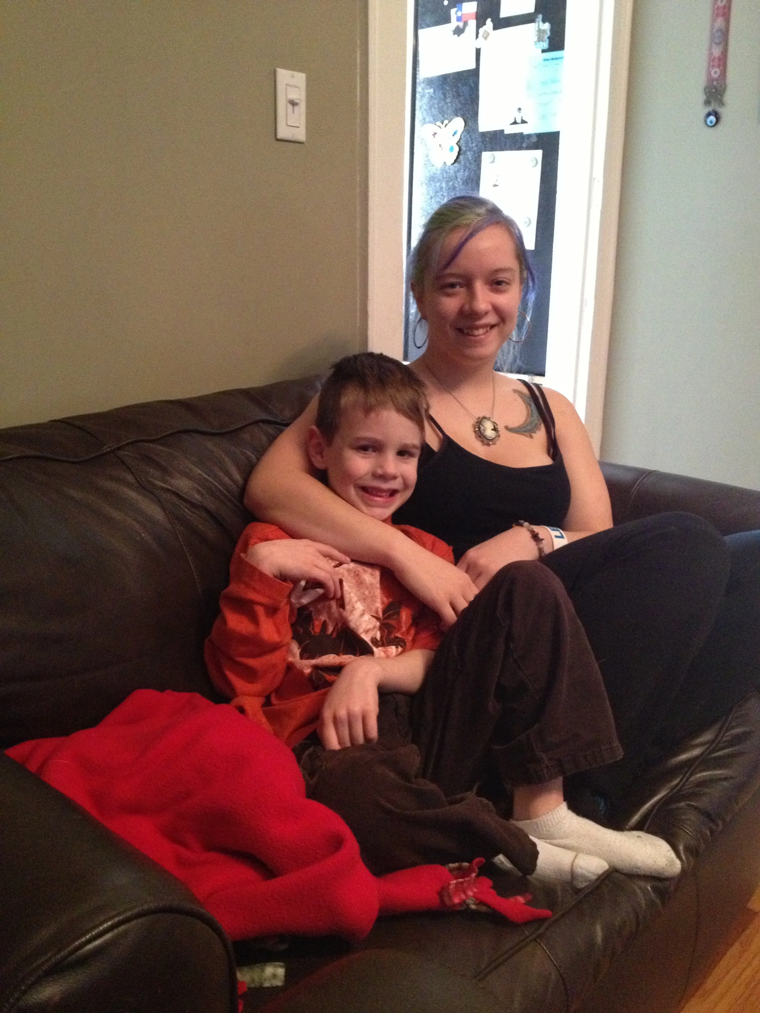 A young woman and a young boy sitting on a dark brown leather couch. The woman has light skin, colorful hair, by her arm around the boy, smiling at the camera. The boy has light skin, short brown hair, wearing an orange and black shirt, smiling at th