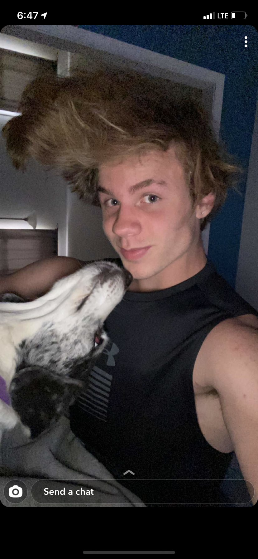 A person with styled hair holding a black and white dog, taking a selfie indoors.