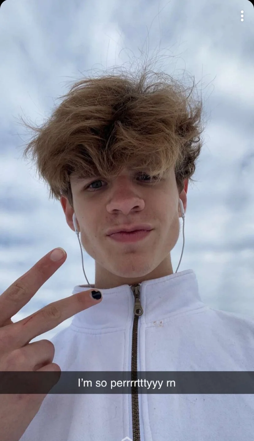 Person with brown hair wearing white earbuds and a white jacket, showing a peace sign with black nail polish on one finger. Text overlay says, 'I'm so perrrrtttyyy rn.'