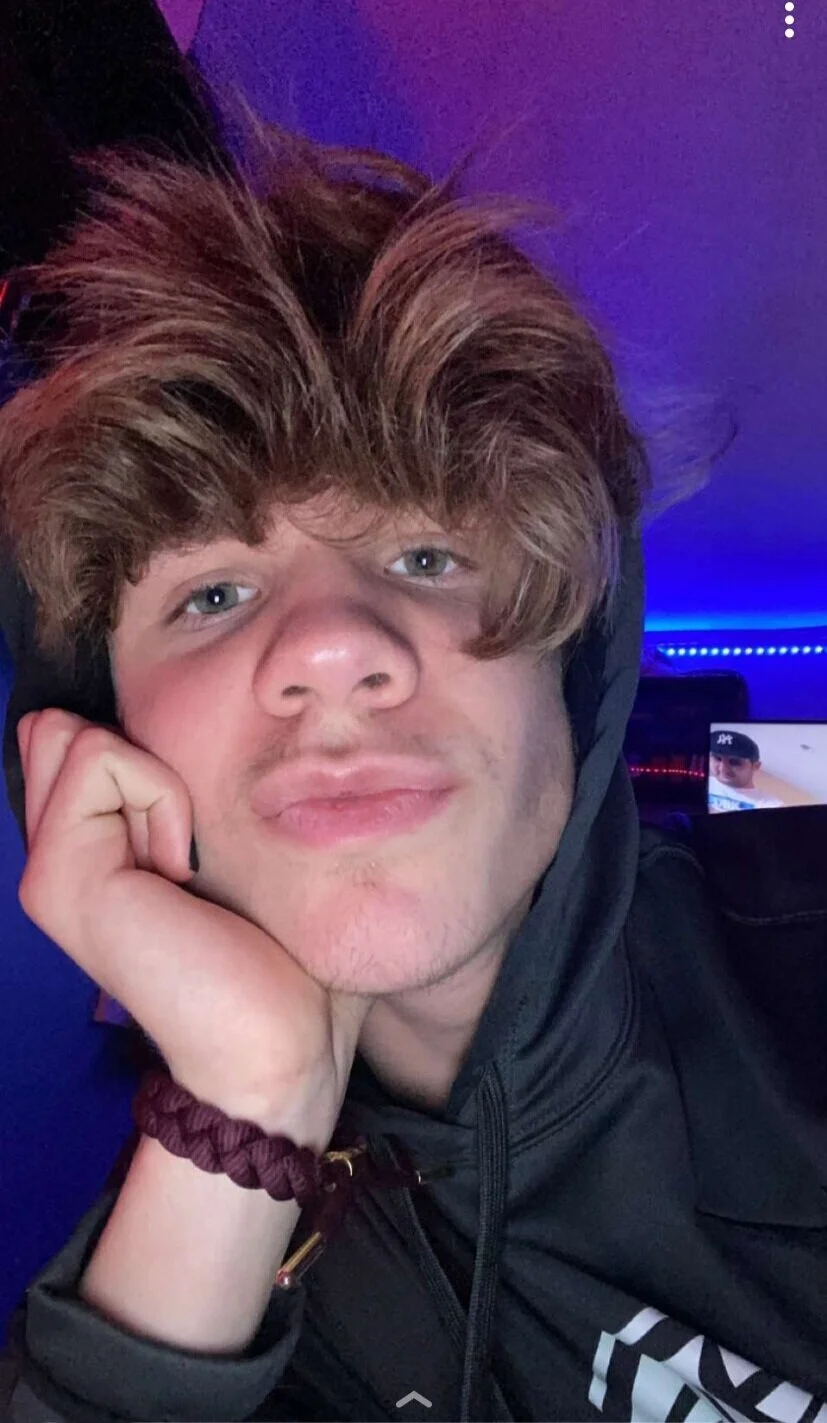 Young person with tousled hair wearing a hoodie, resting their head on their hand, in a room with blue lighting.