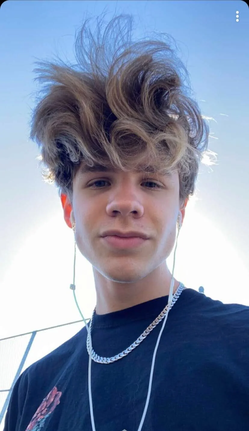 Person with tousled hair, wearing a black shirt, chain necklace, and earbuds, in outdoor setting with blue sky background.