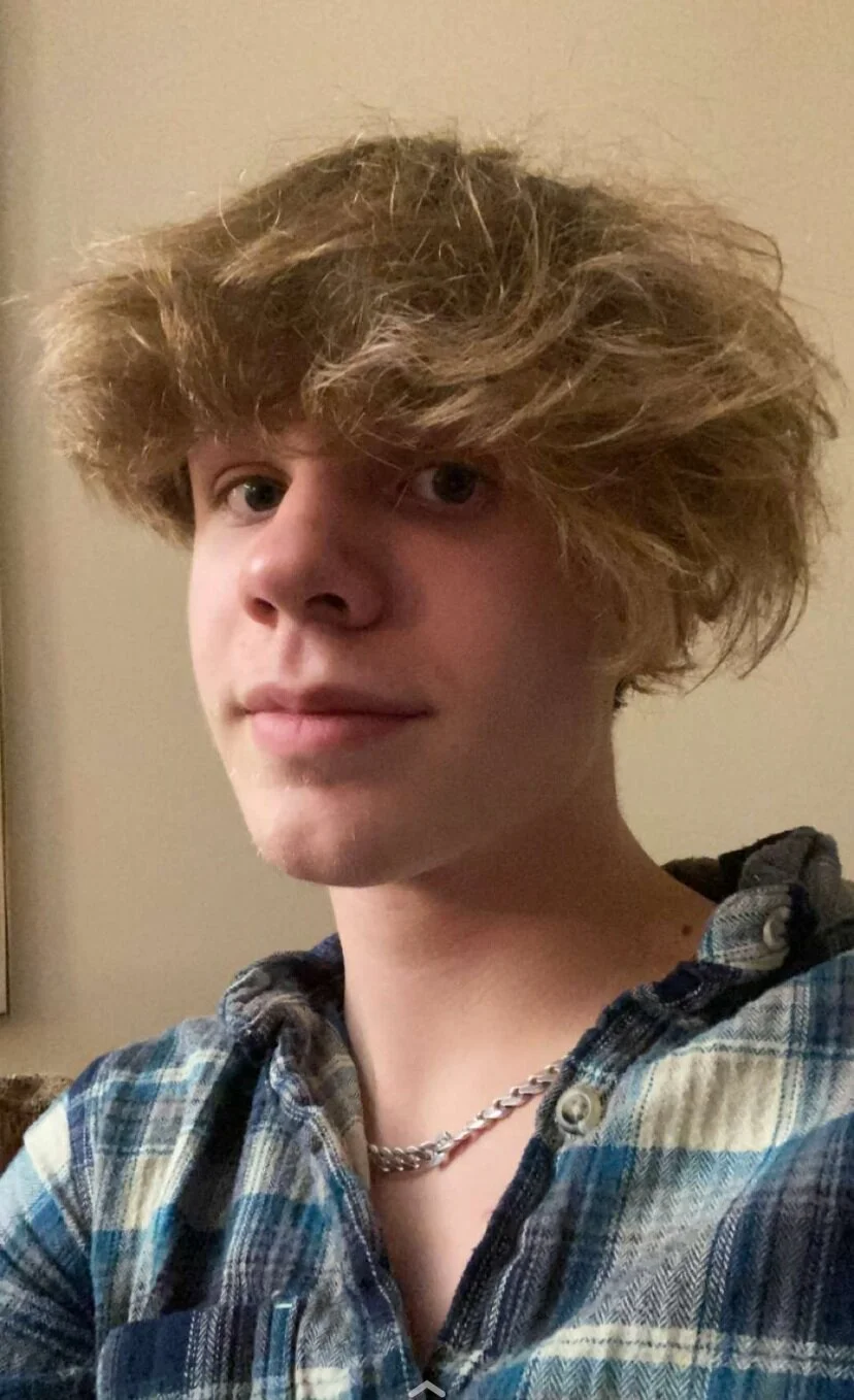 Person with tousled hair wearing a plaid shirt and chain necklace.