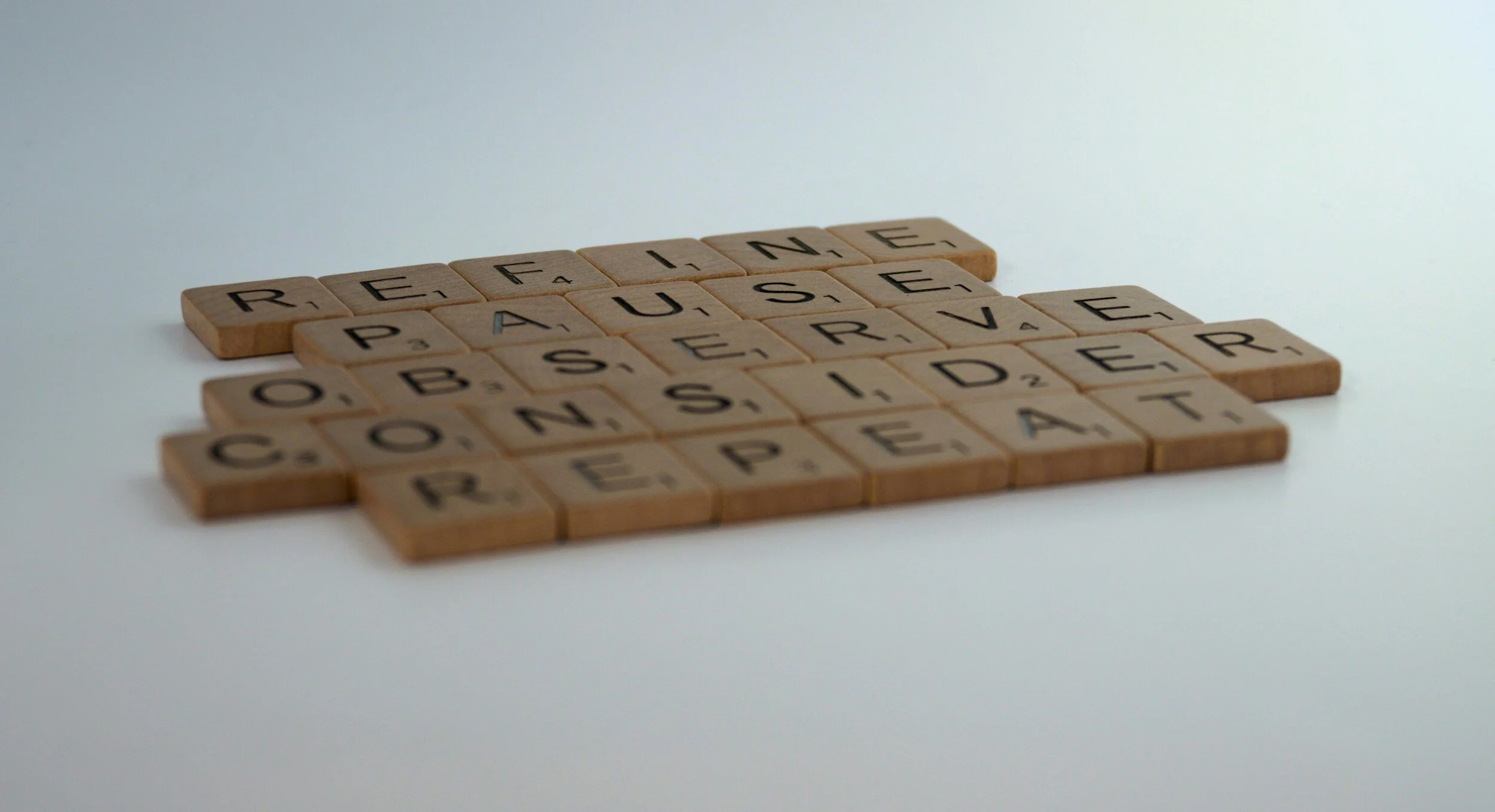 Wooden letter tiles spelling 'REFINE, PAUSE, OBSERVE, CONSIDER, REPEAT' on a white surface.