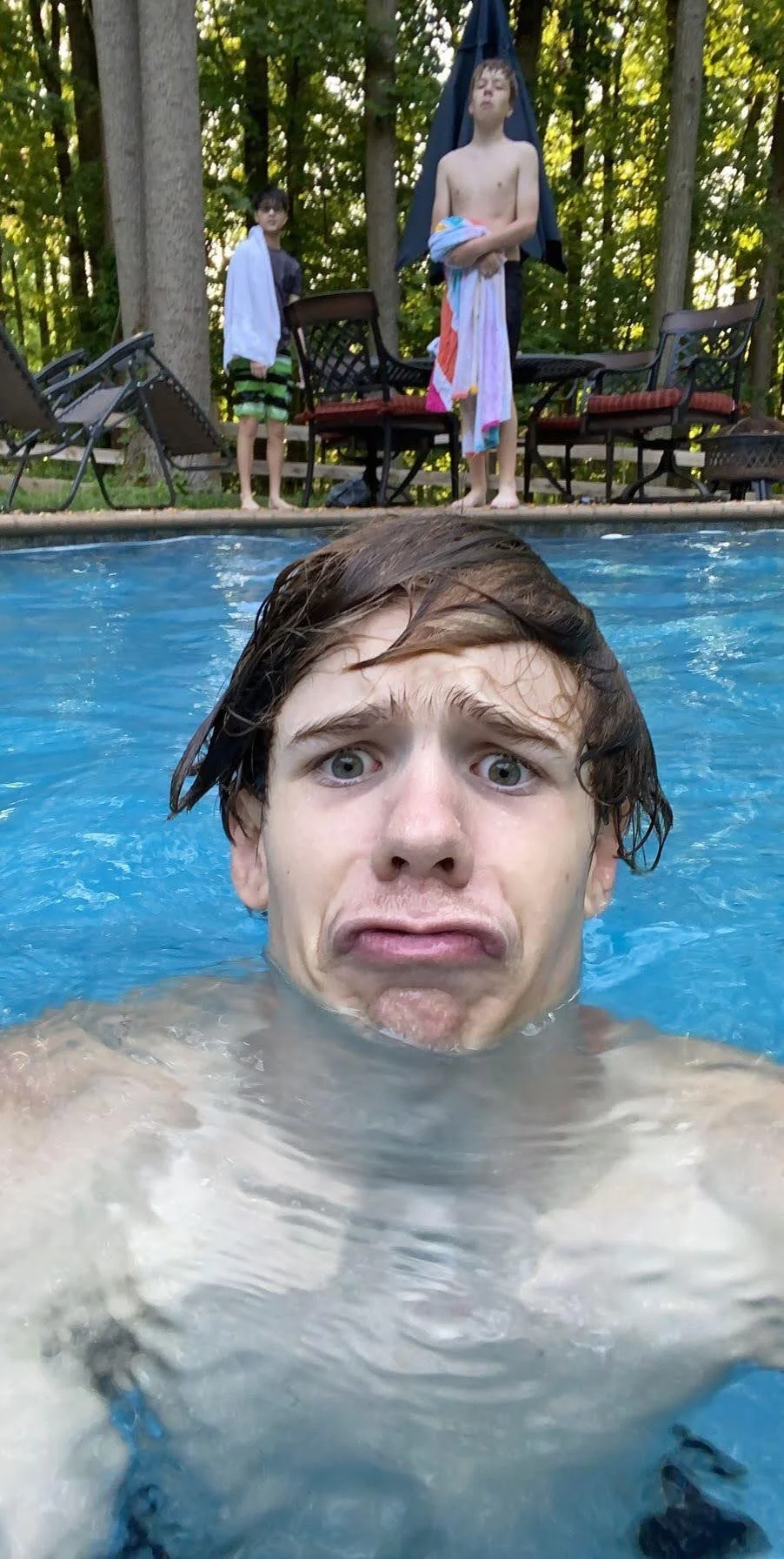 A person making a funny face while partially submerged in a swimming pool, with two other people in the background by the poolside holding towels, surrounded by trees and outdoor furniture.