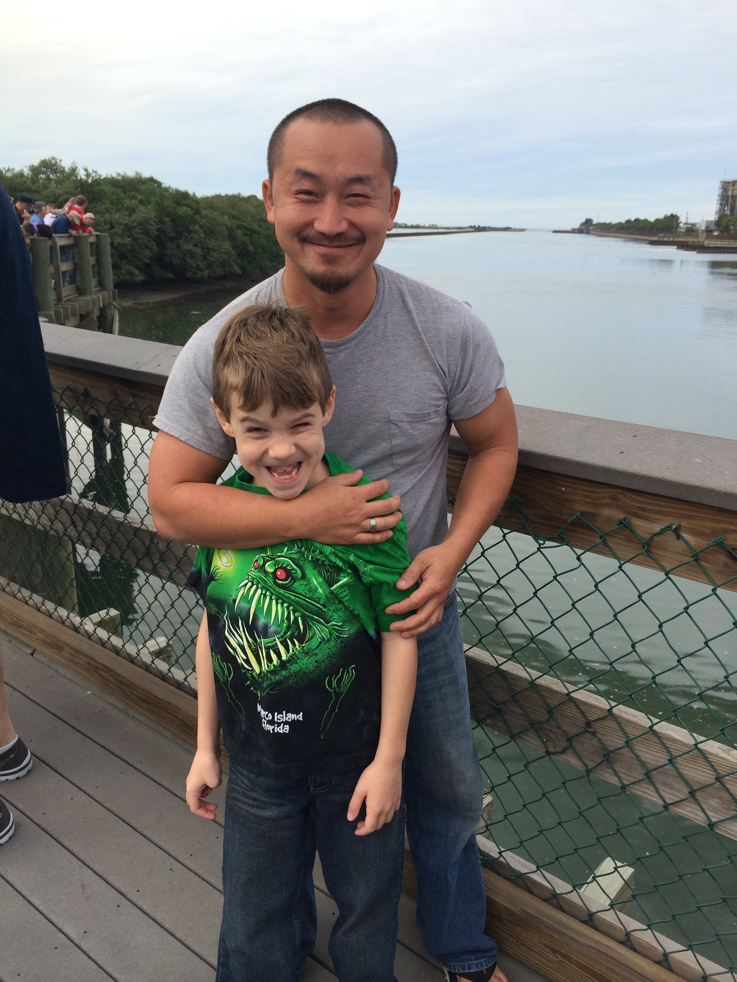 A man and a young boy standing on a wooden pier by the water, hugging and smiling. The boy is wearing a green T-shirt with a dinosaur graphic, and the man is wearing a gray T-shirt.