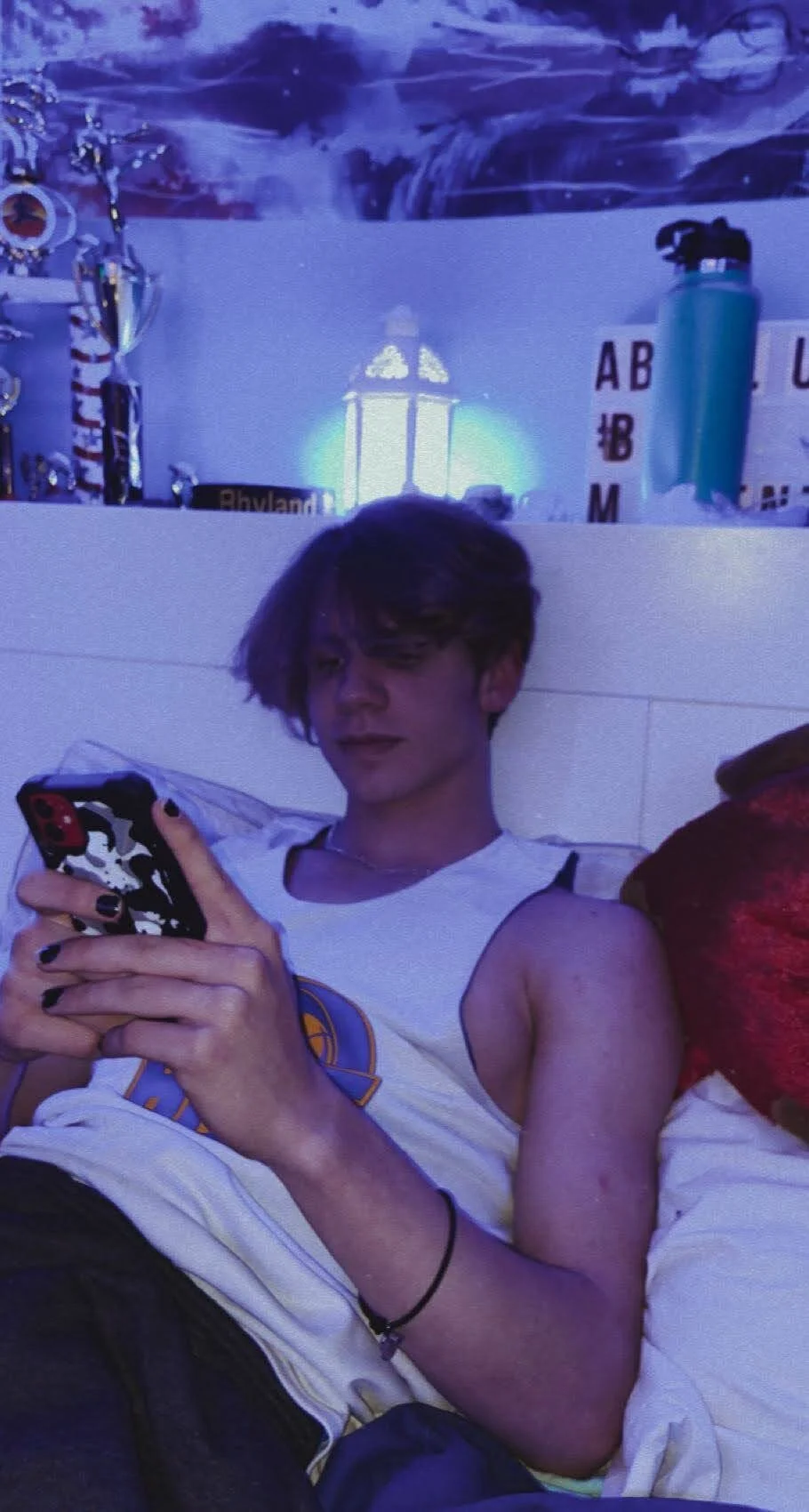 Person with painted nails using smartphone while lying on bed, trophies and decorative items in the background.