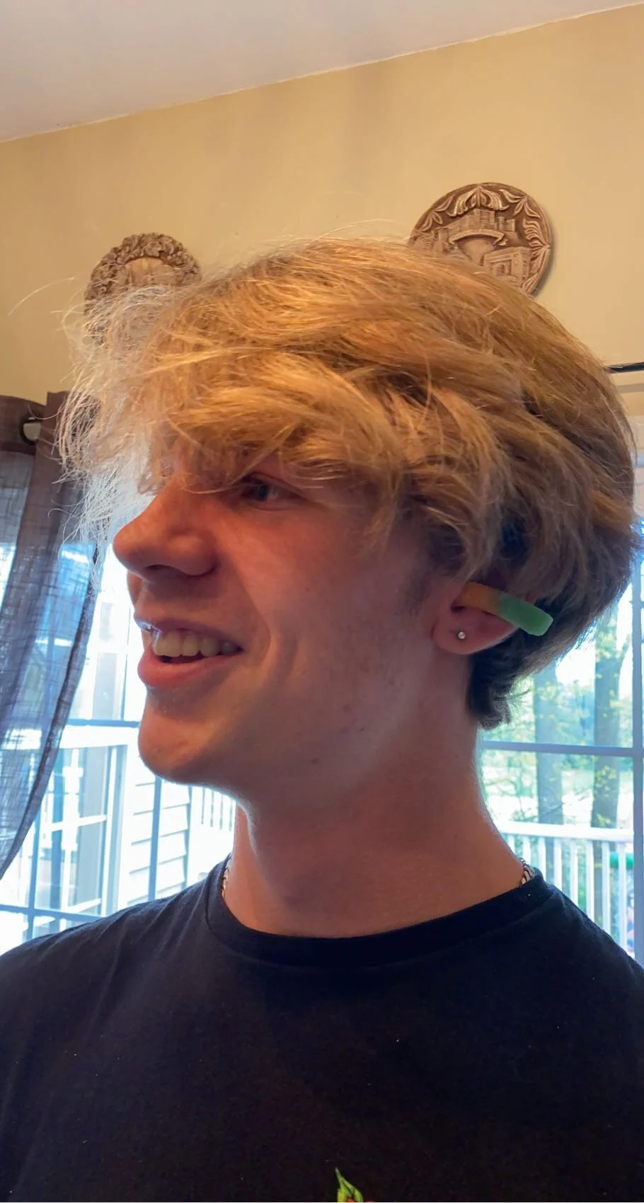 A young person with blonde hair wearing a black shirt, a green gummy candy on their ear, smiling in a room with a window and decorative wall hangings.
