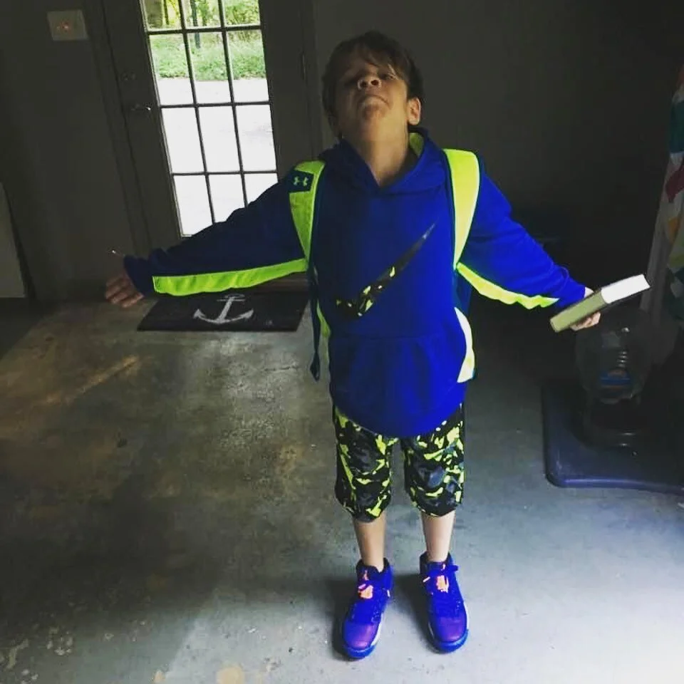 A young boy standing with arms outstretched inside a room, wearing a blue hoodie, neon yellow and black shorts, and purple sneakers. He is holding a book in his right hand and has a backpack on.