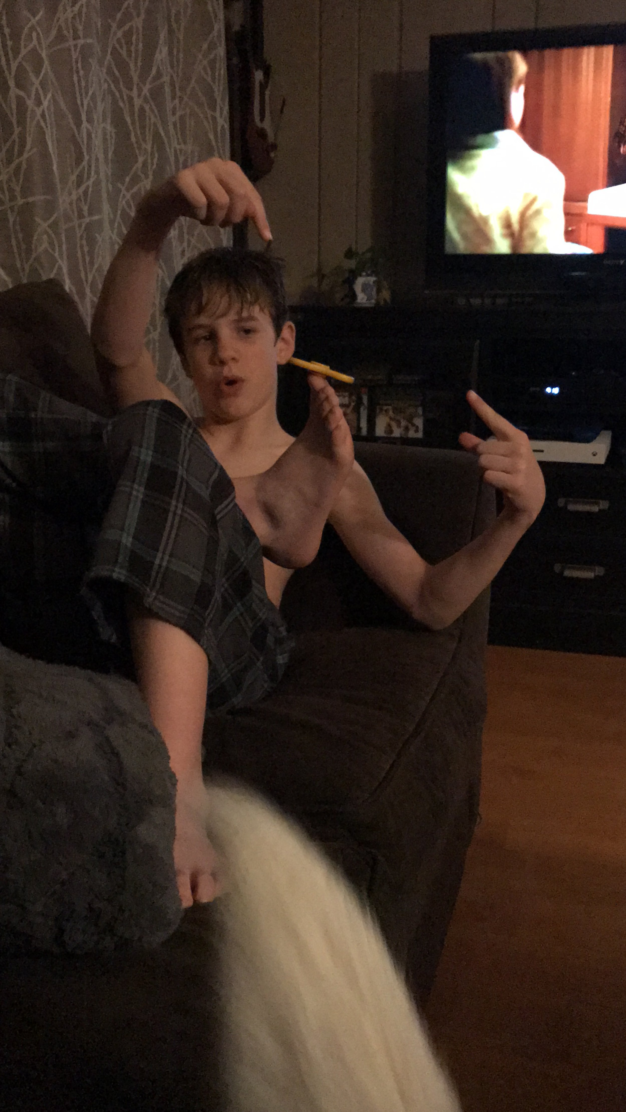 A young boy sitting on a couch making a playful gesture with his hands, shirtless, with a TV in the background showing a person wearing a yellow shirt.