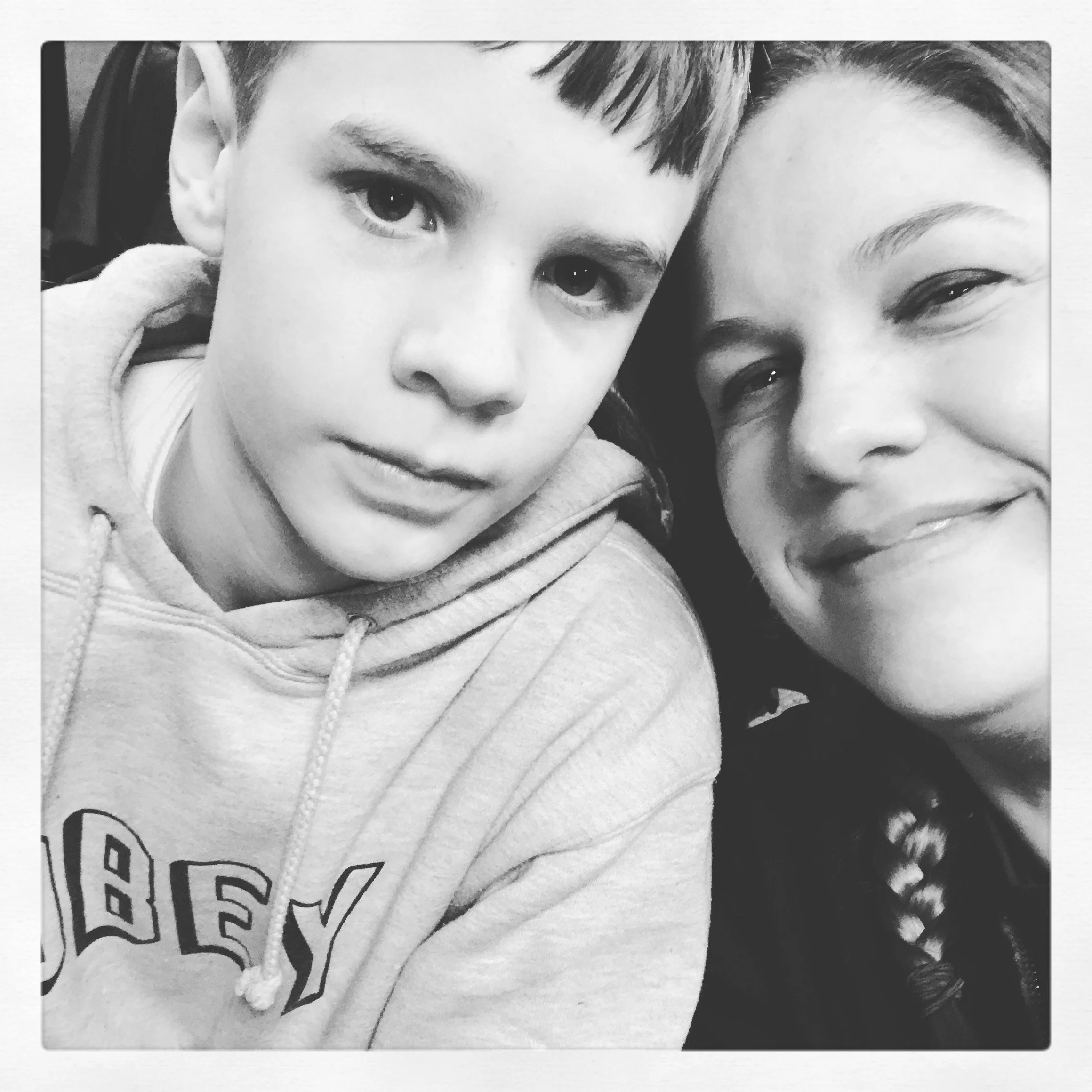 A black and white photo of a boy and a woman smiling and taking a selfie together. The boy is wearing a hoodie, and the woman has curly hair.