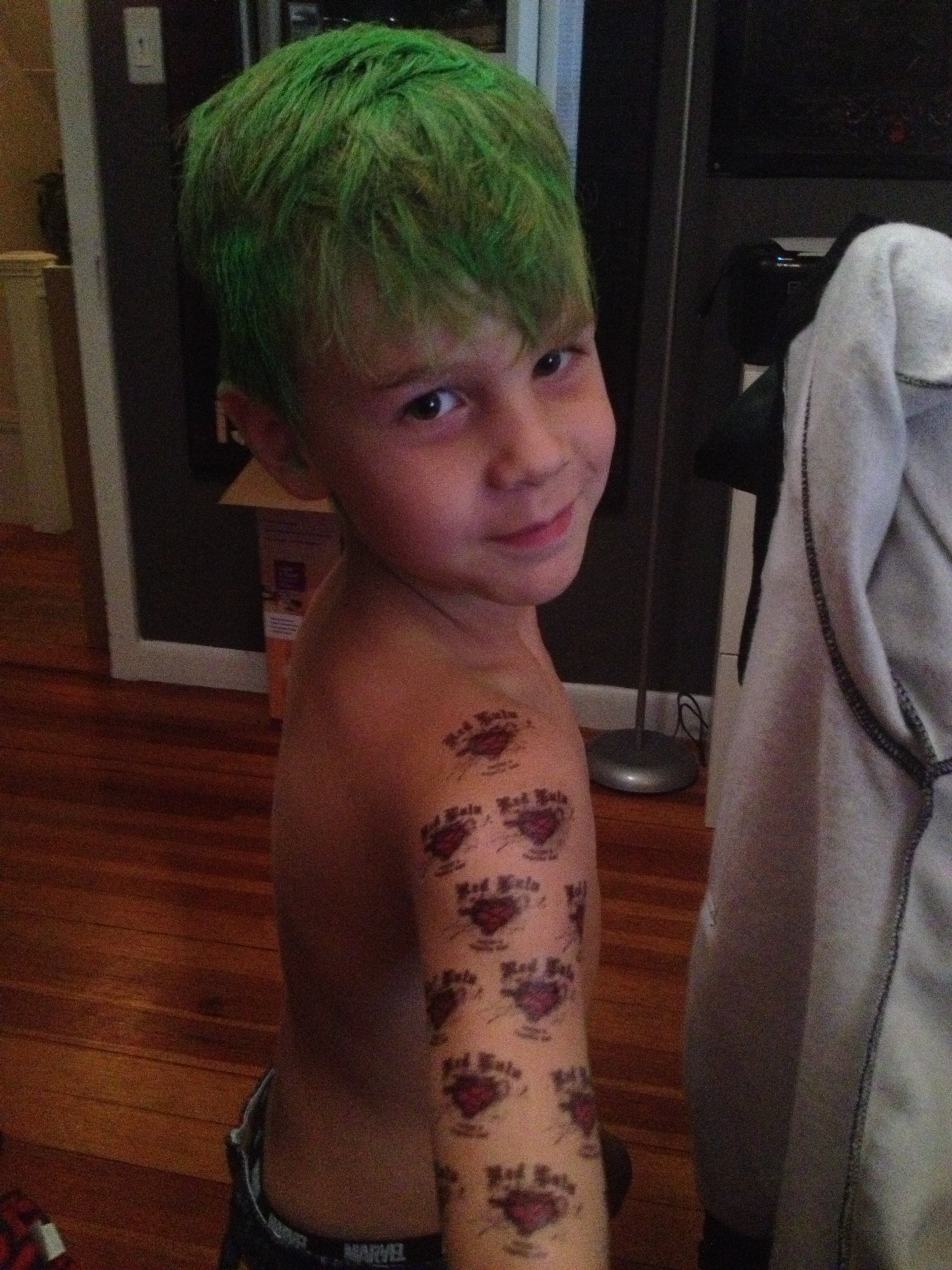 A young boy with short hair dyed green, smiling and looking at the camera, with temporary tattoos on his left arm and shoulder, in a room with hardwood floors and various household items.