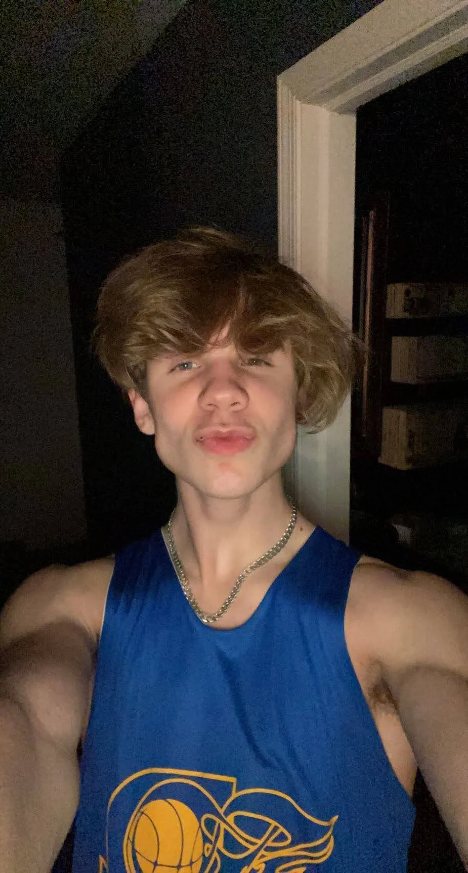 Person in a blue tank top with basketball graphic posing in a dimly lit room.