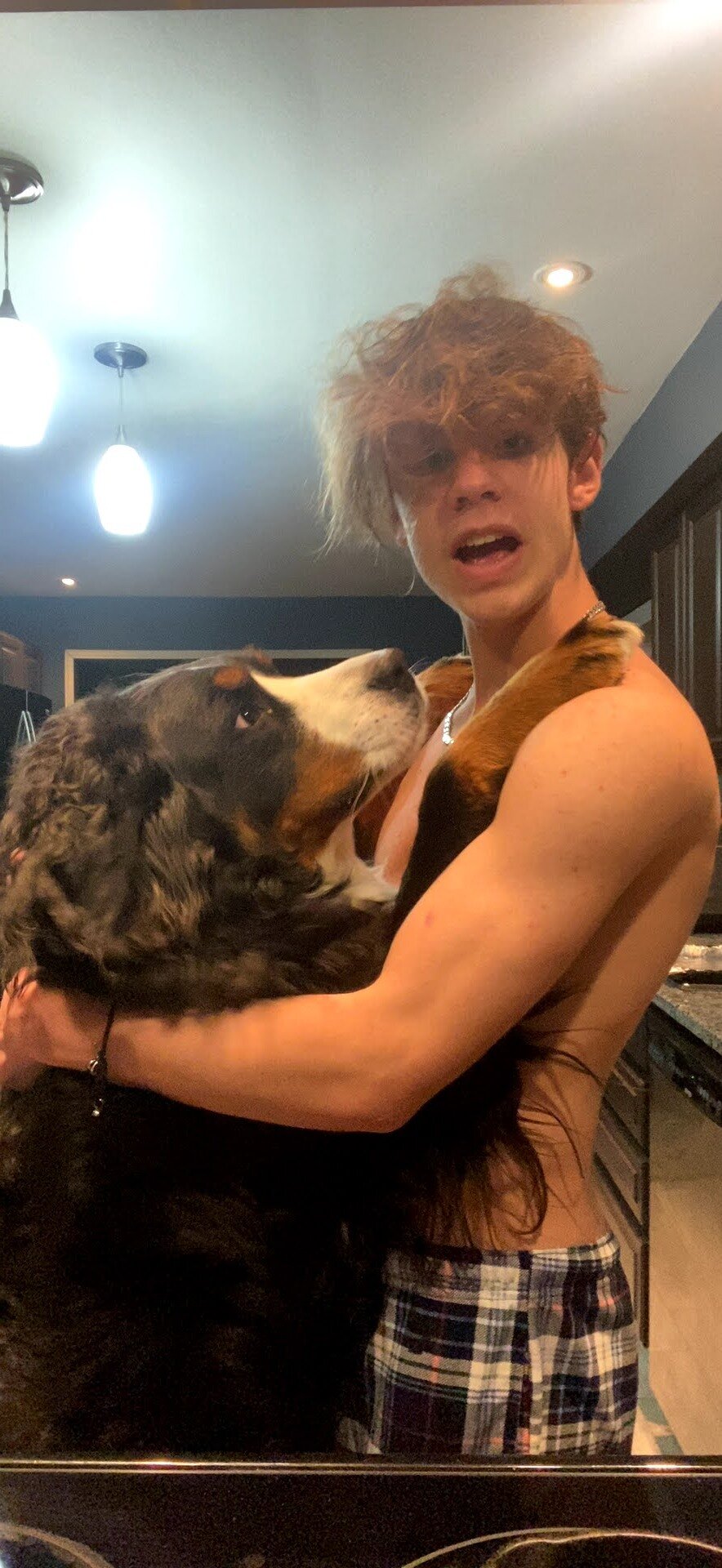 A young man with messy blonde hair holding a Bernese mountain dog in a kitchen.