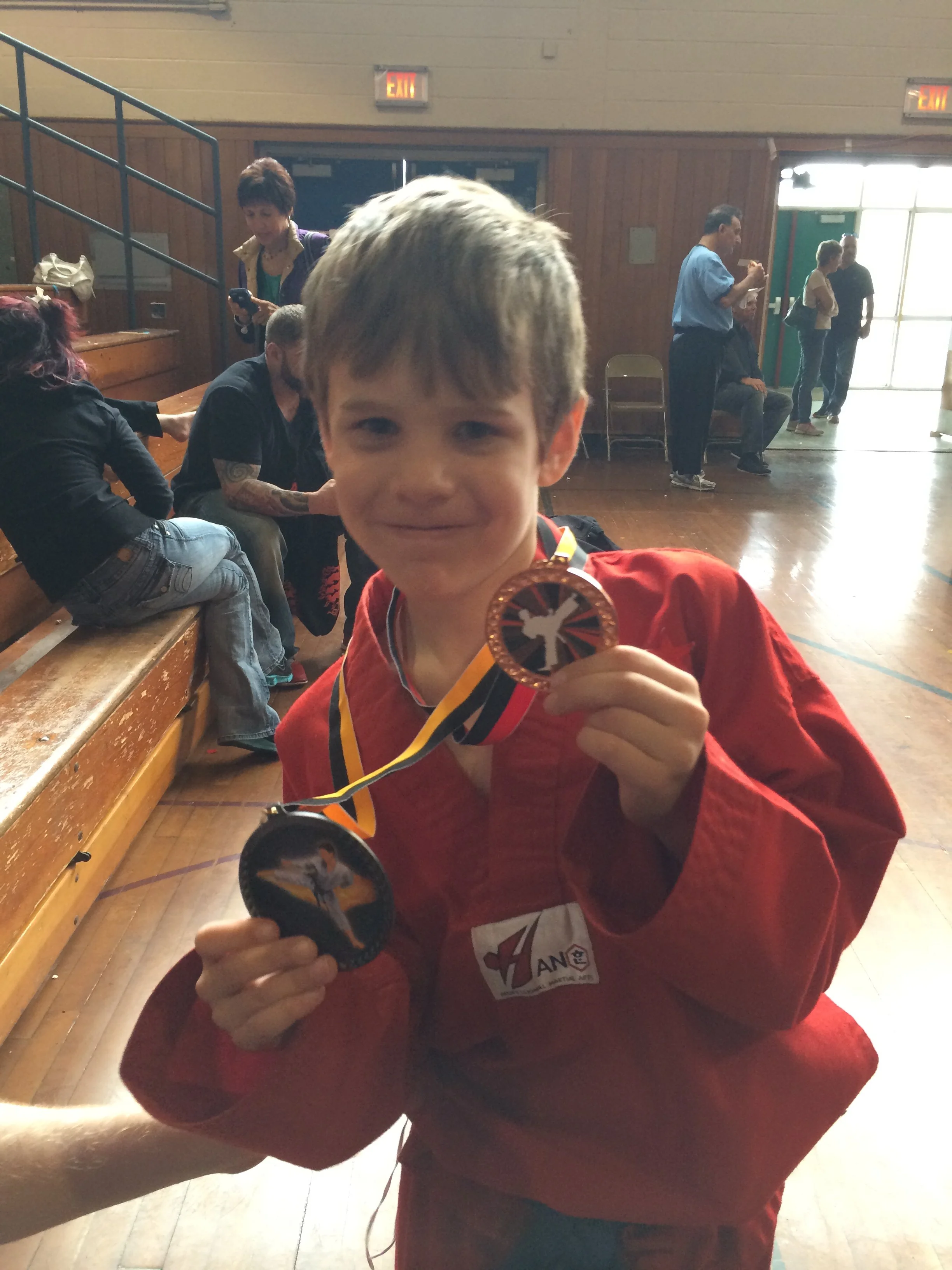 Young boy in a red martial arts uniform holding up two medals inside a gymnasium.