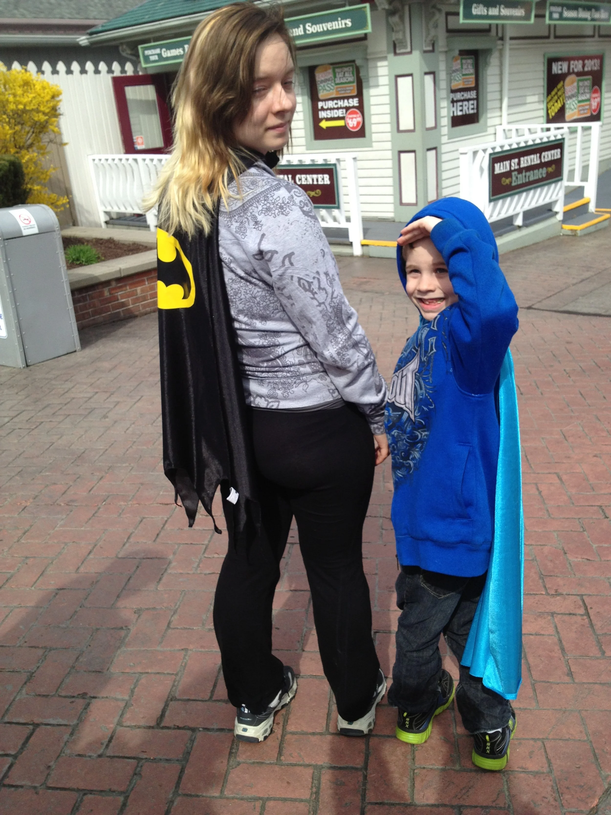 A young woman dressed as Batman and a boy dressed as a superhero standing on a brick sidewalk in front of a gift shop, with signs and an entrance in the background.
