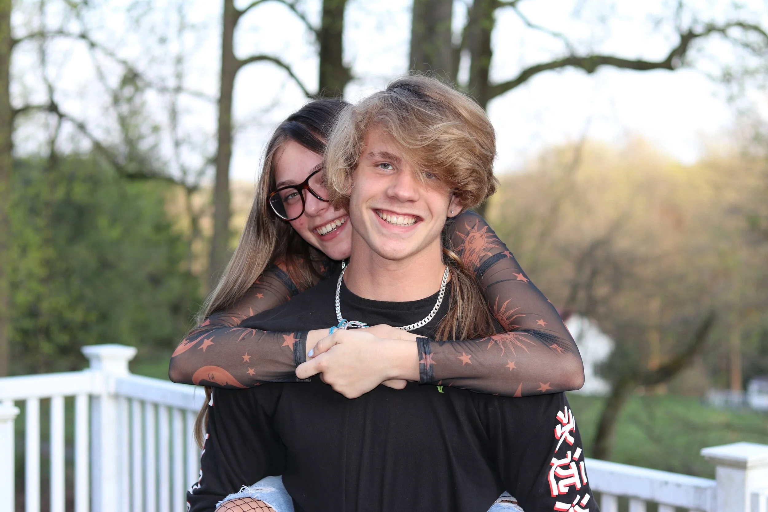 A young couple outdoors, the girl wearing glasses and smiling with her arms around the boy's shoulders.