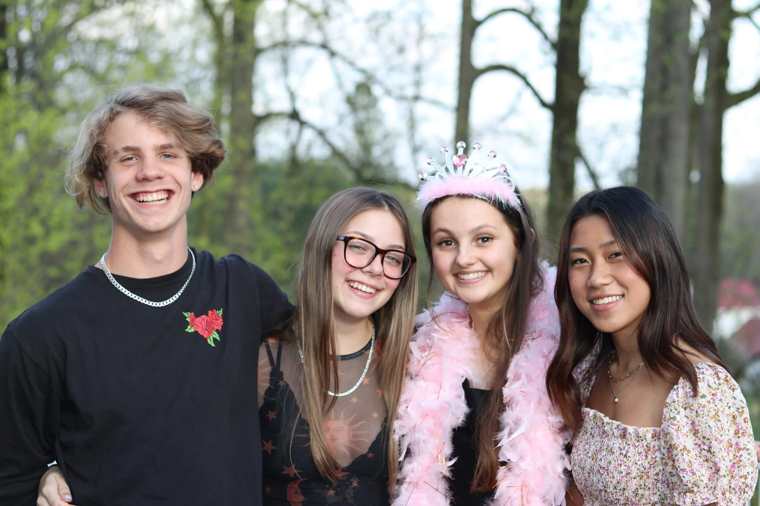 Four smiling teenagers standing together outdoors, with one wearing a tiara and pink feather boa.