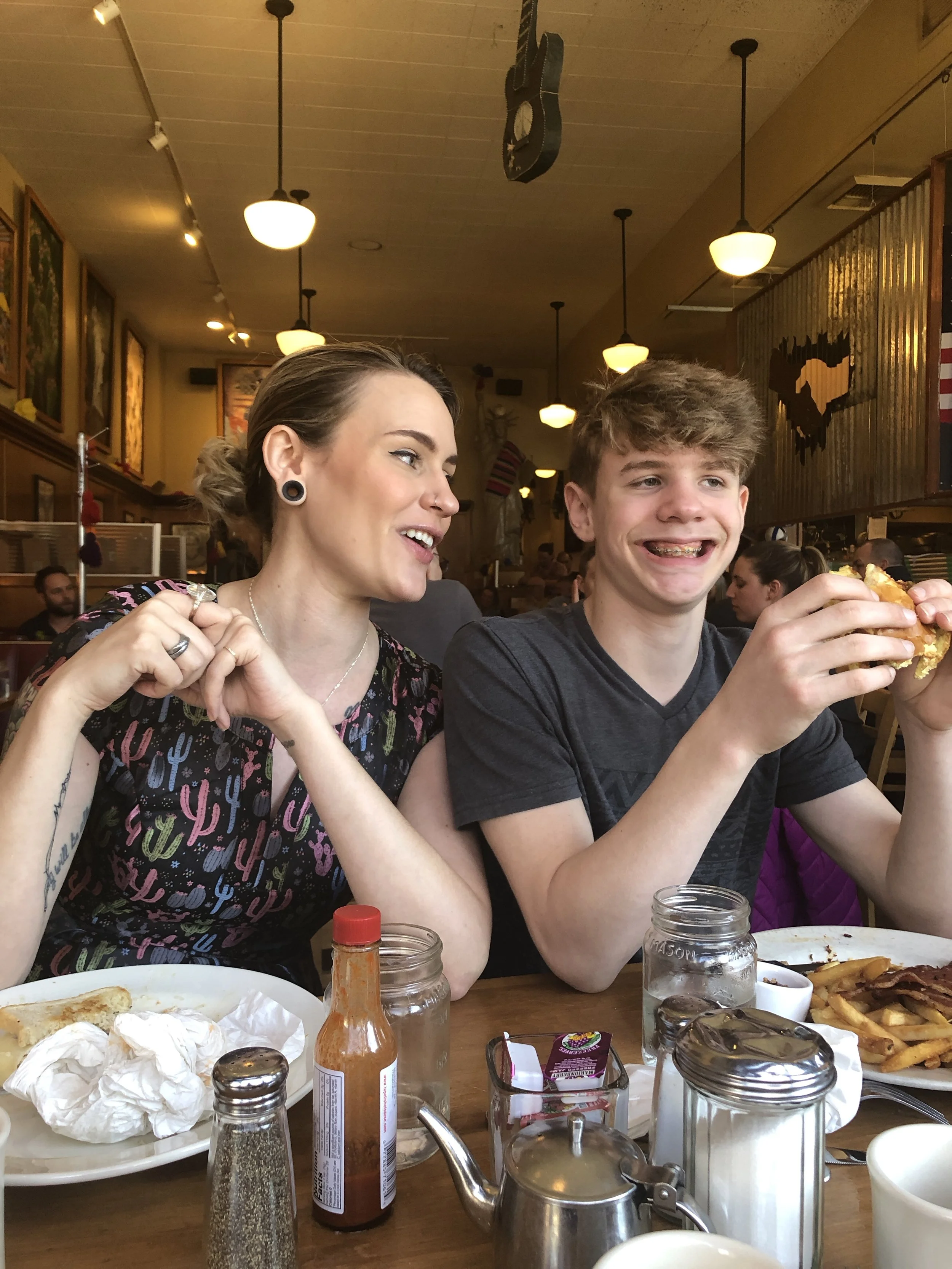 A woman and a young man laughing and eating at a restaurant table with food, condiments, and drinks.