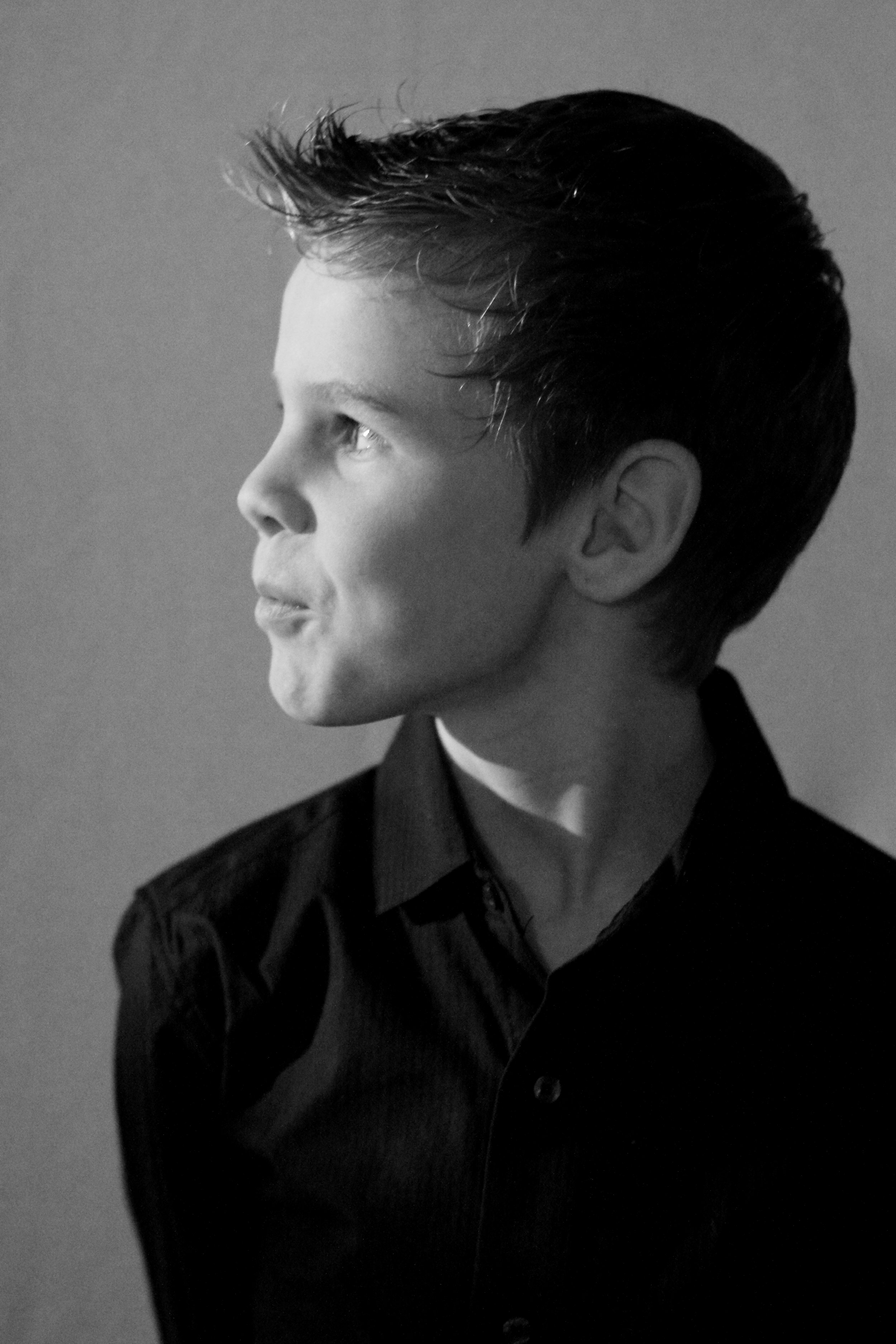 A black and white portrait of a young boy with short hair, looking to the left, wearing a dark collared shirt.