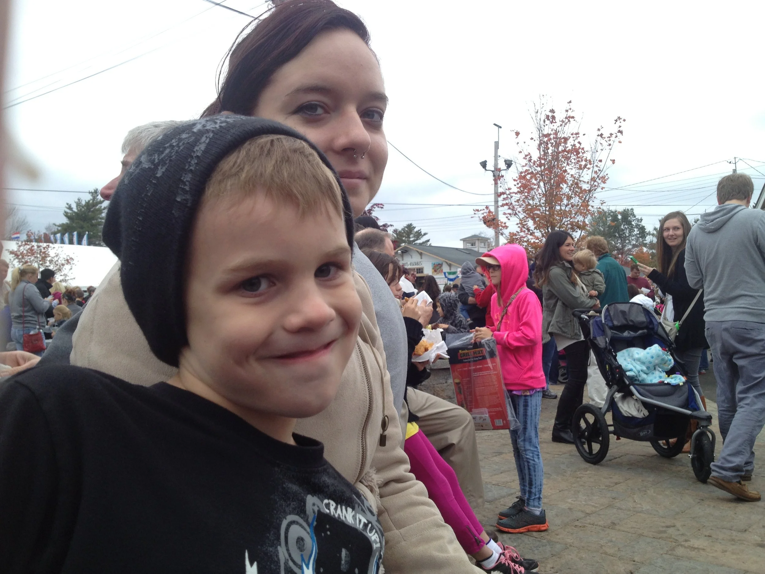A young boy with a black beanie and a girl with dark hair and a piercing, sitting outdoors with a crowd, some pushing strollers, under overcast skies during fall.