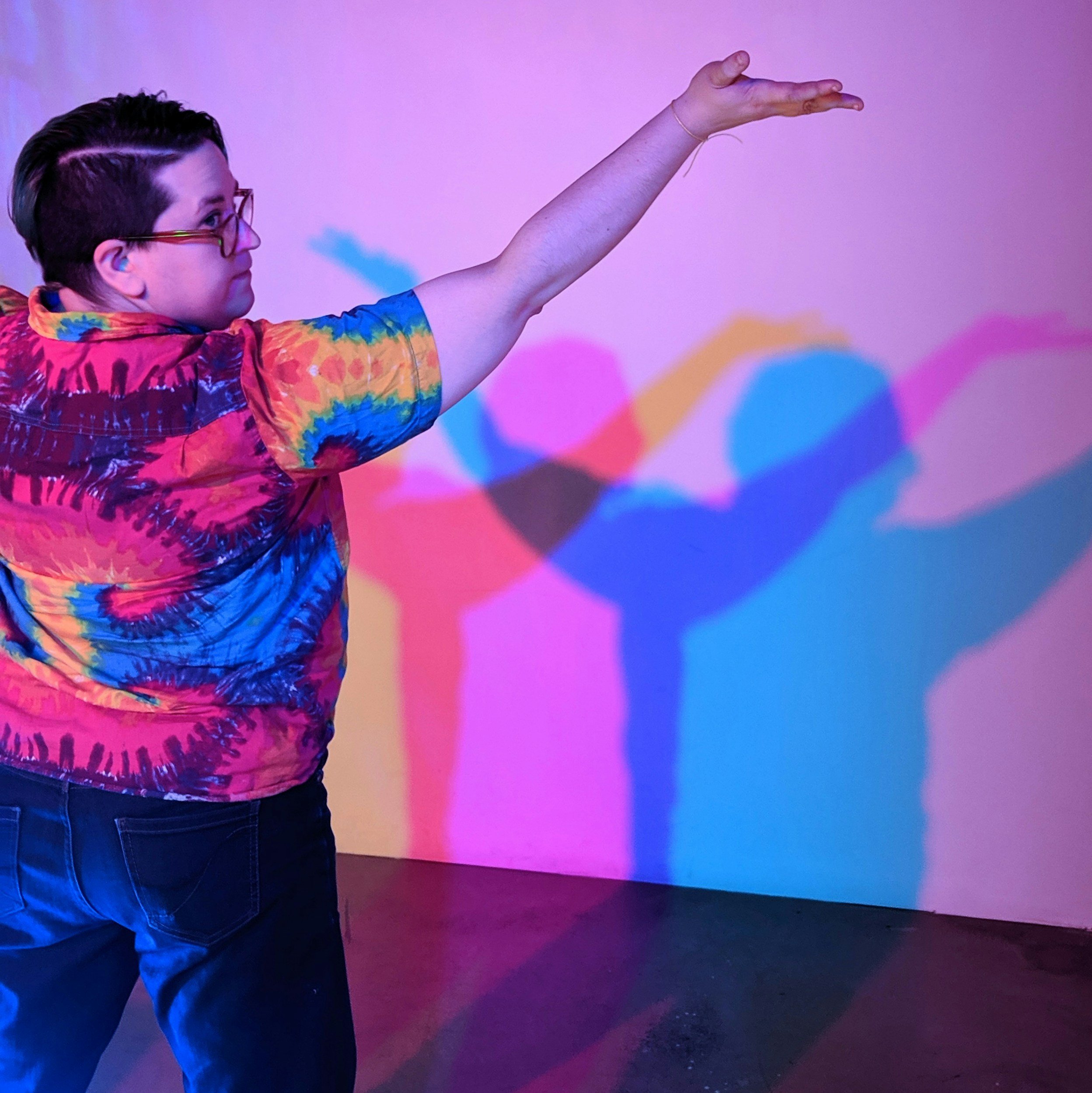 A person wearing a rainbow tie dye shirt with their arms raised over head and their back to the camera, showing expansive movement as a way to explore self expression in therapy.