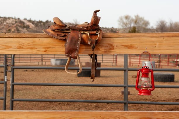 A leather saddle hanging on a wooden rail with a red lantern hanging nearby, in an outdoor riding arena.