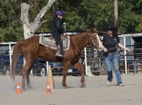 A young girl in helmet riding a horse, guided by a woman in a hat, at an outdoor riding arena with orange cones and trees in the background.
