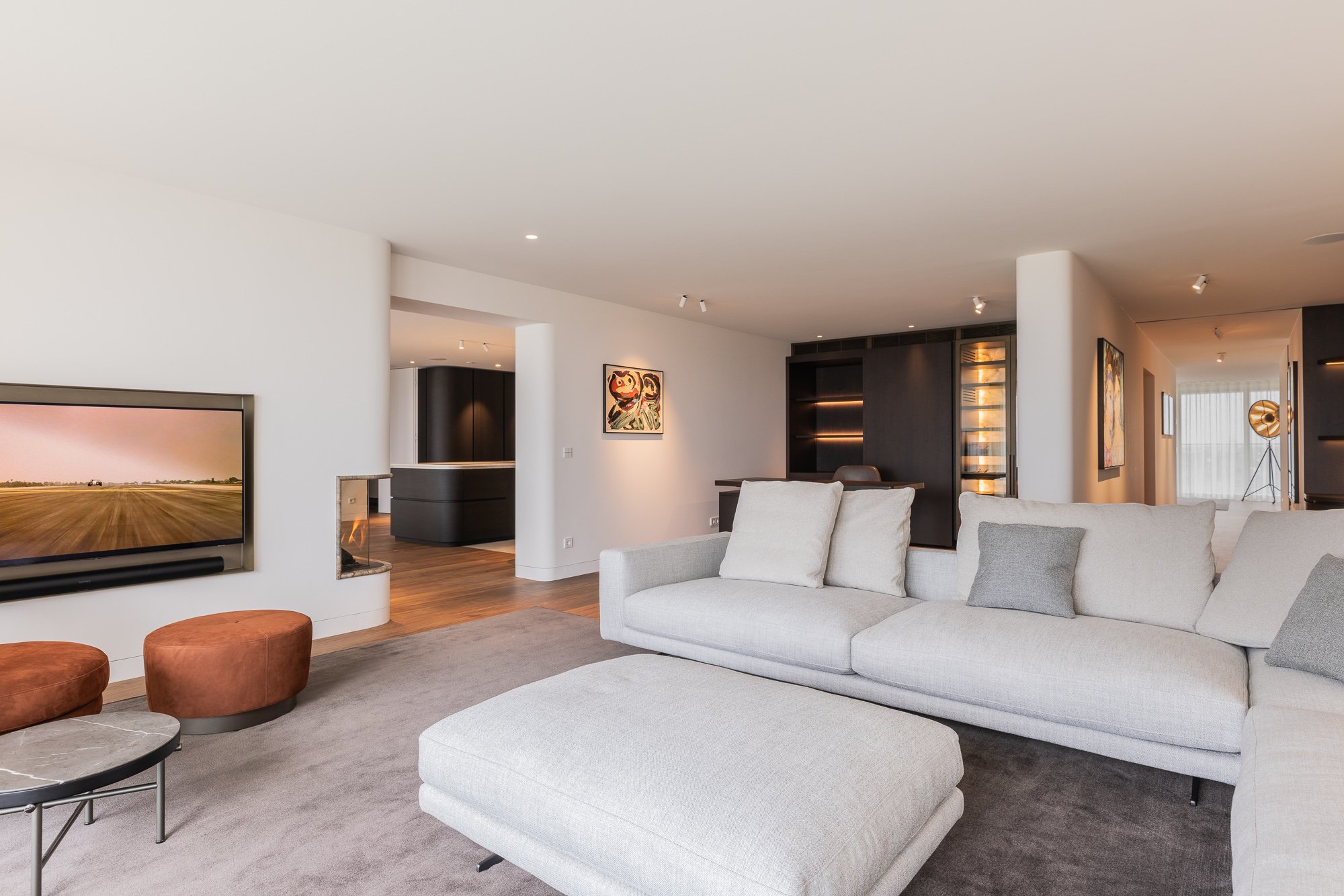 Moderne, ruime woonkamer met lichtgrijze hoekbank, grote tv, open haard, kunstwerken en zwarte inbouwkasten.